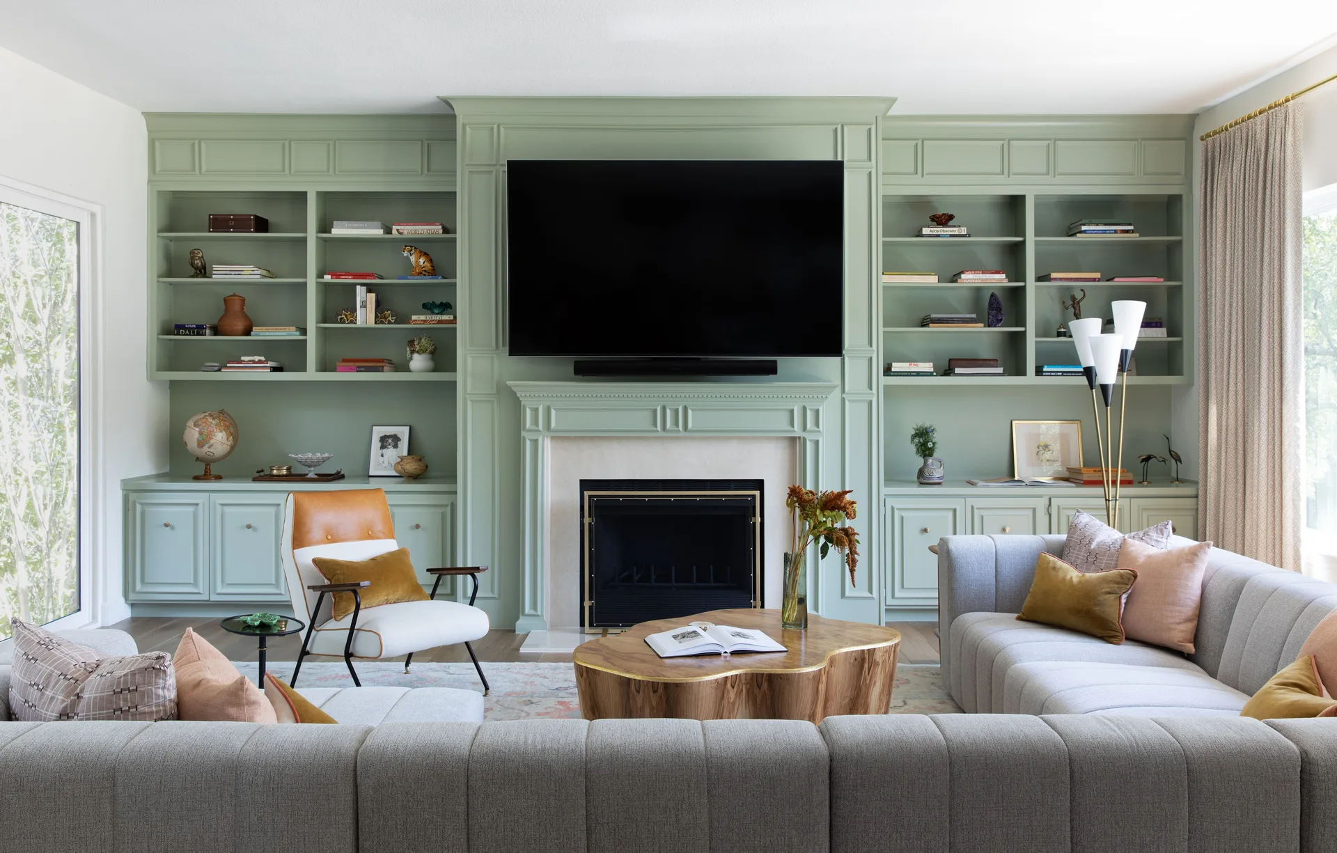 A transitional living room featuring a custom wall of sage green built-in bookshelves, a gray sectional sofa, and an organic wood coffee table.
