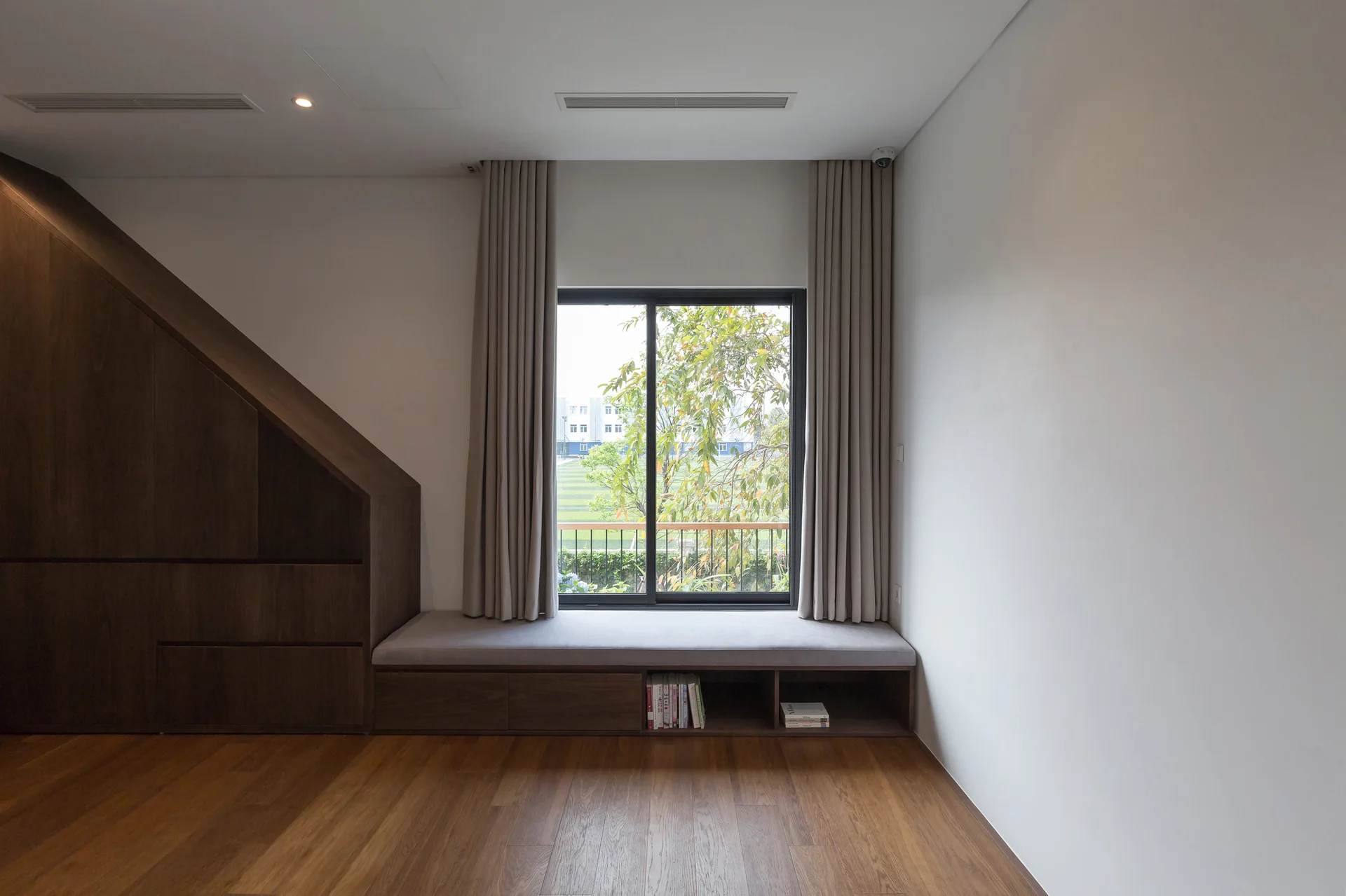 A minimalist room features a dark wood storage cabinet with a slanted top alongside a cushioned window seat with long beige curtains.