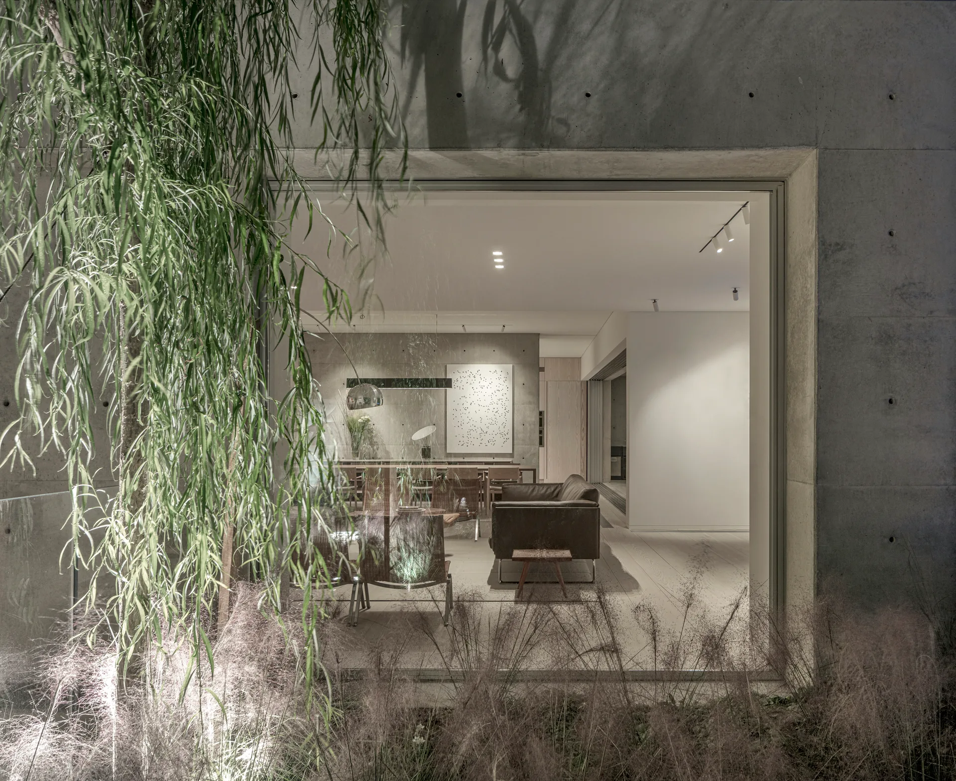 A warm modern living space is viewed through a large square window set into a raw concrete exterior wall flanked by soft green foliage.