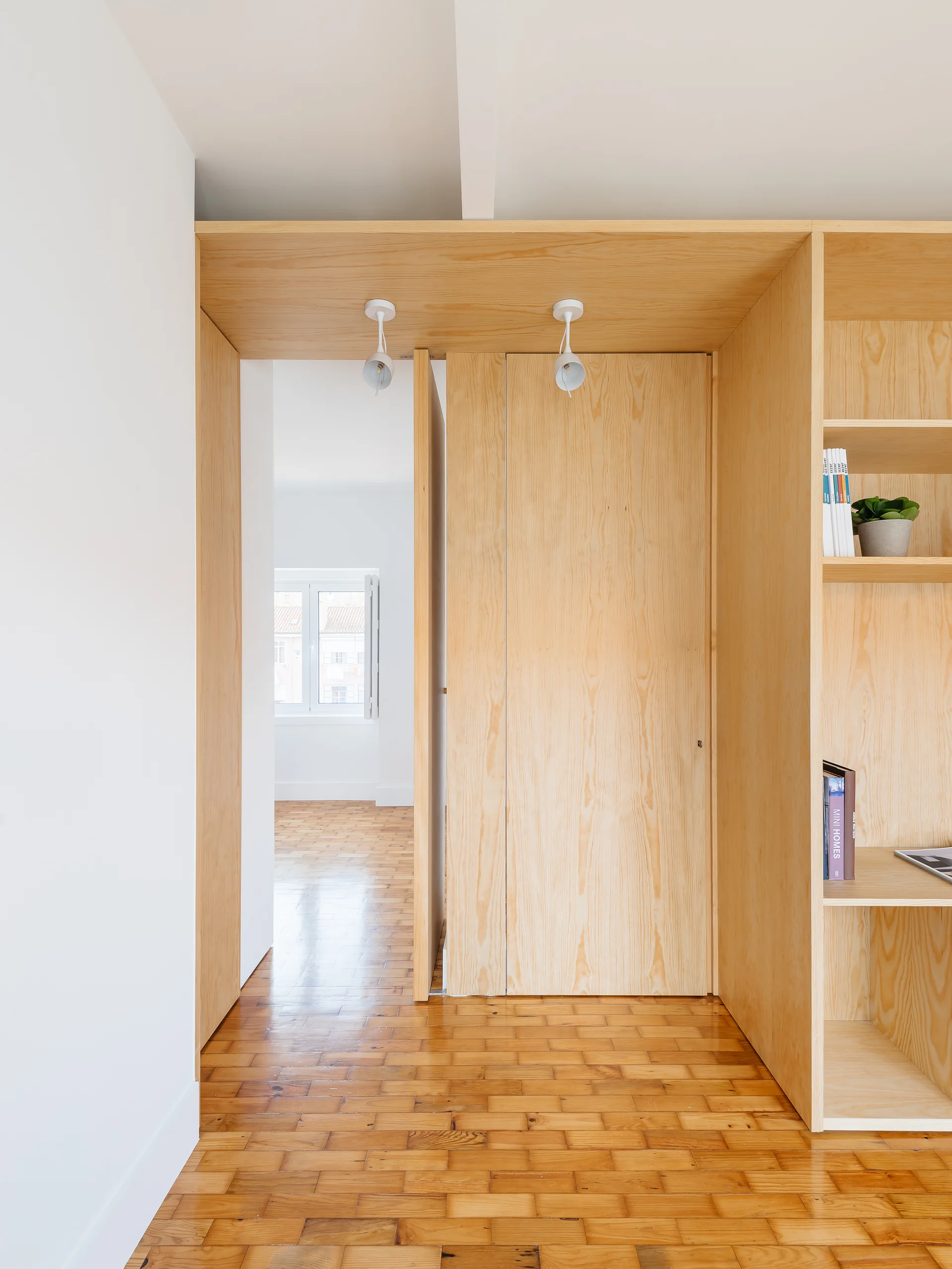 A minimalist hallway featuring custom light plywood built-in cabinetry, white walls, ceiling spotlights, and warm parquet wood flooring.