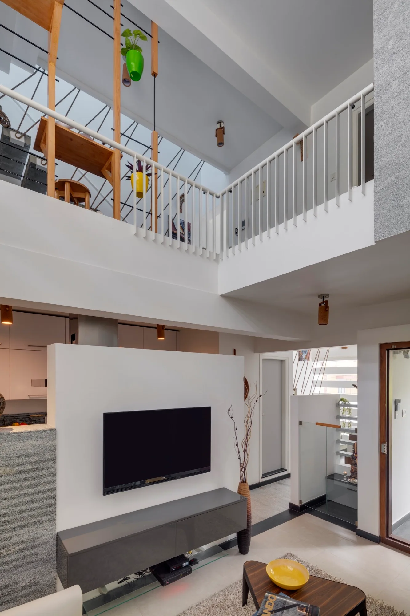 A bright double-height living room features a white partition wall with a mounted television, a grey media console, and a mezzanine level above.