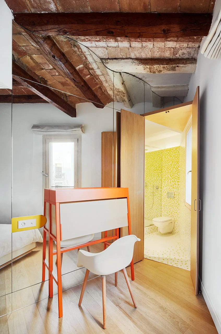 A modern bedroom features a bright orange desk next to a mirrored wall, with a brick vaulted ceiling above and an adjacent yellow tiled bathroom visible.