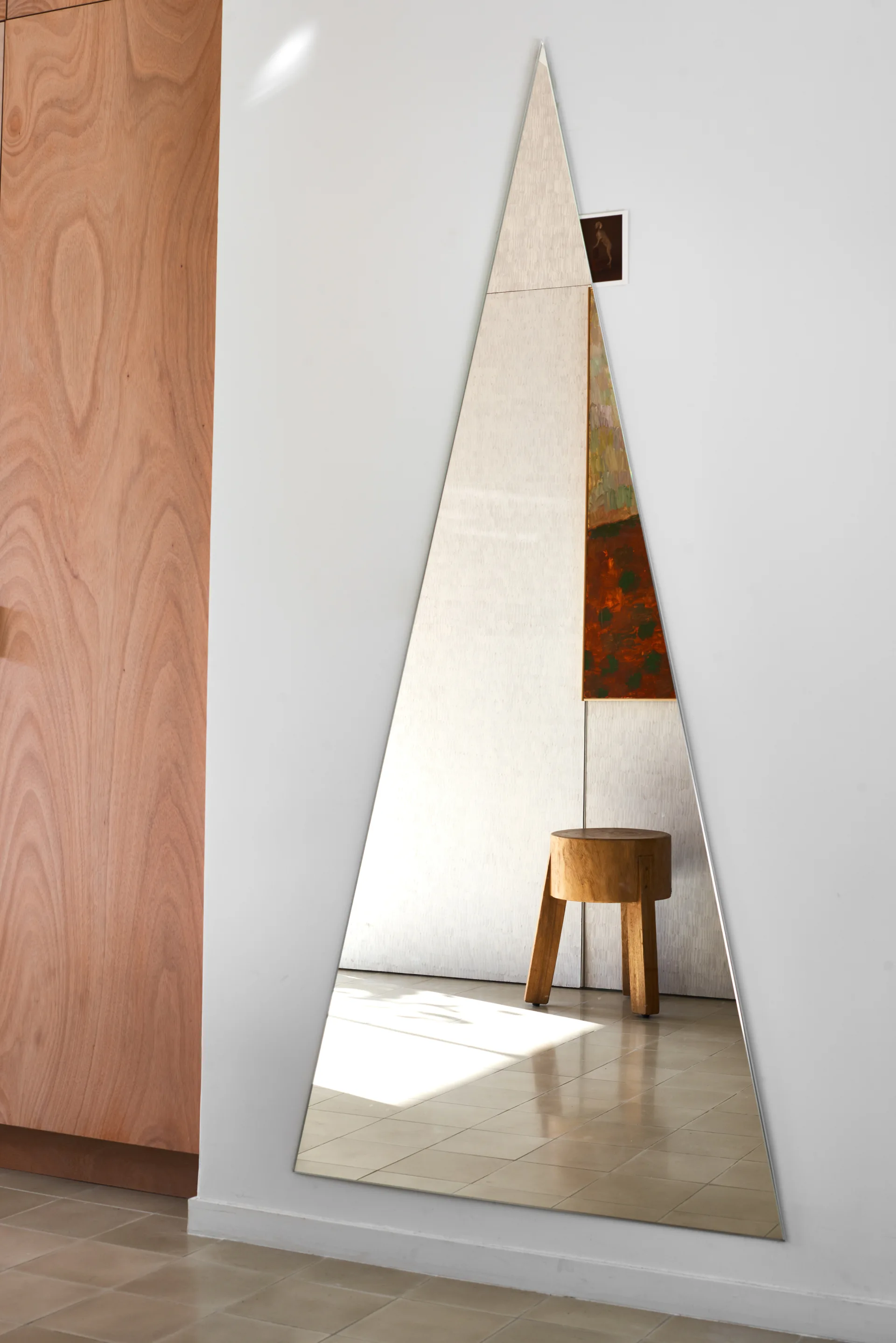 A tall triangular mirror mounted on a white wall next to wood paneling reflects a small three-legged wooden stool.