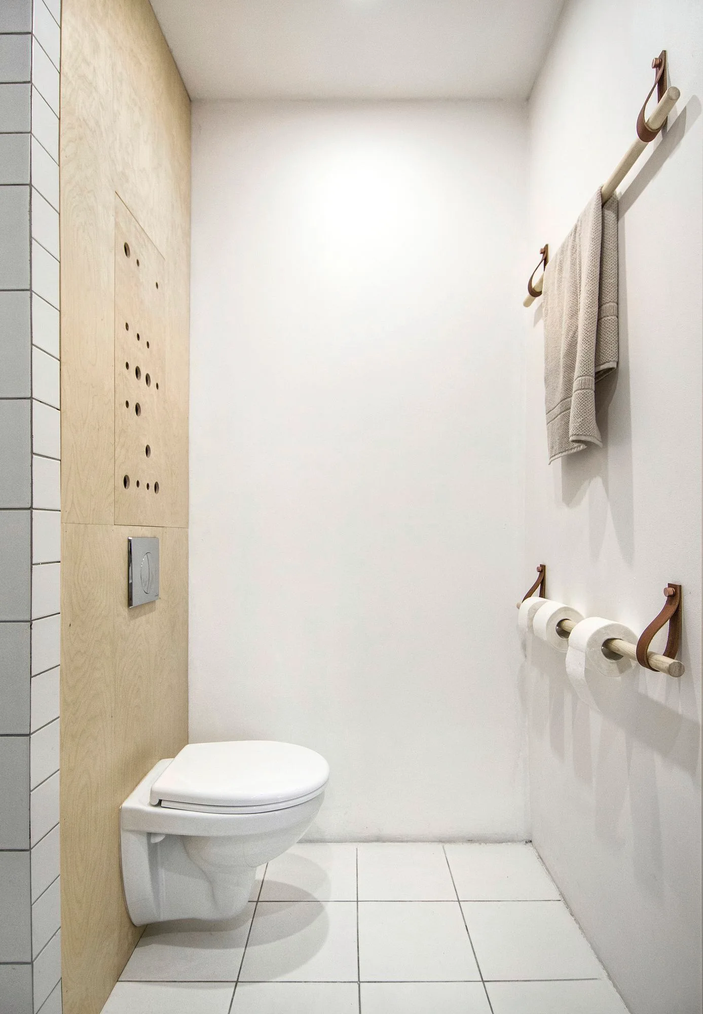 A minimalist powder room with white grid tiled floors, a wall-mounted toilet, and wooden dowel holders suspended by leather straps.