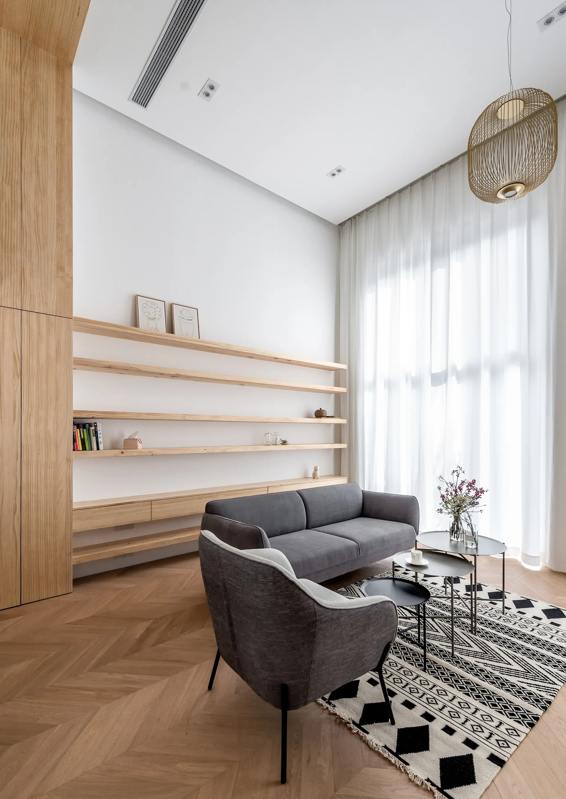 A minimalist living room features a grey sofa and armchair, long light wood floating shelves, and herringbone flooring.