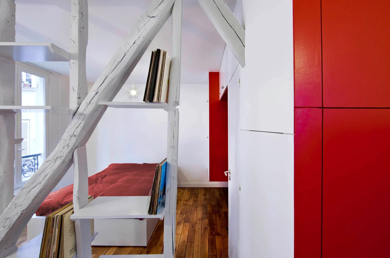 A loft bedroom featuring a white painted timber support frame used as shelving next to a red platform bed and a red wardrobe cabinet.
