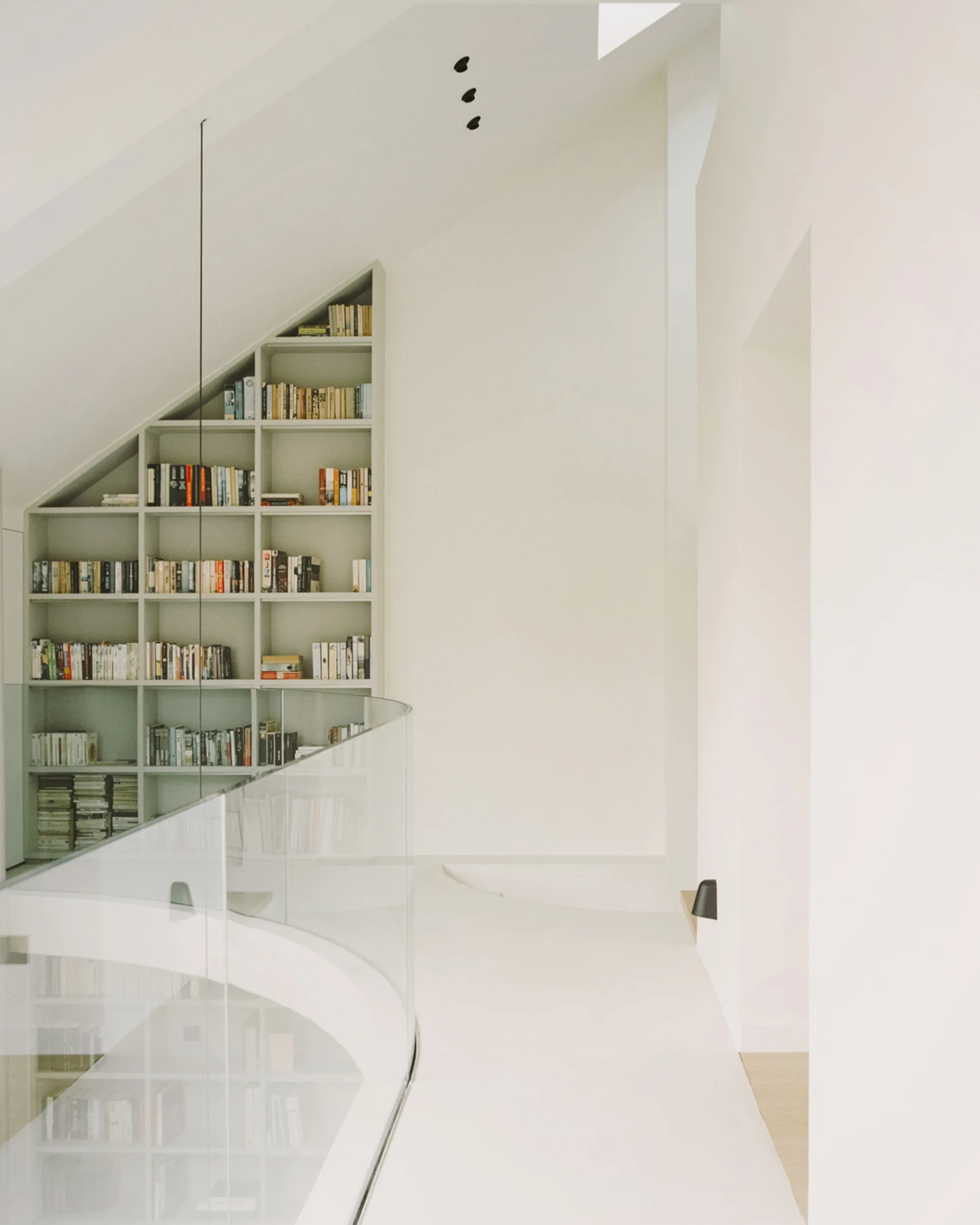 A minimalist off-white landing features a custom slanted bookshelf under an angled ceiling and a curved glass railing.