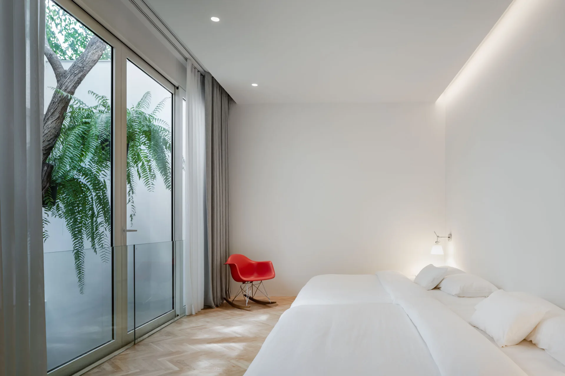 A minimalist white bedroom features a bed with white sheets, a red rocking chair, herringbone wood floors, and a large window looking into a courtyard.