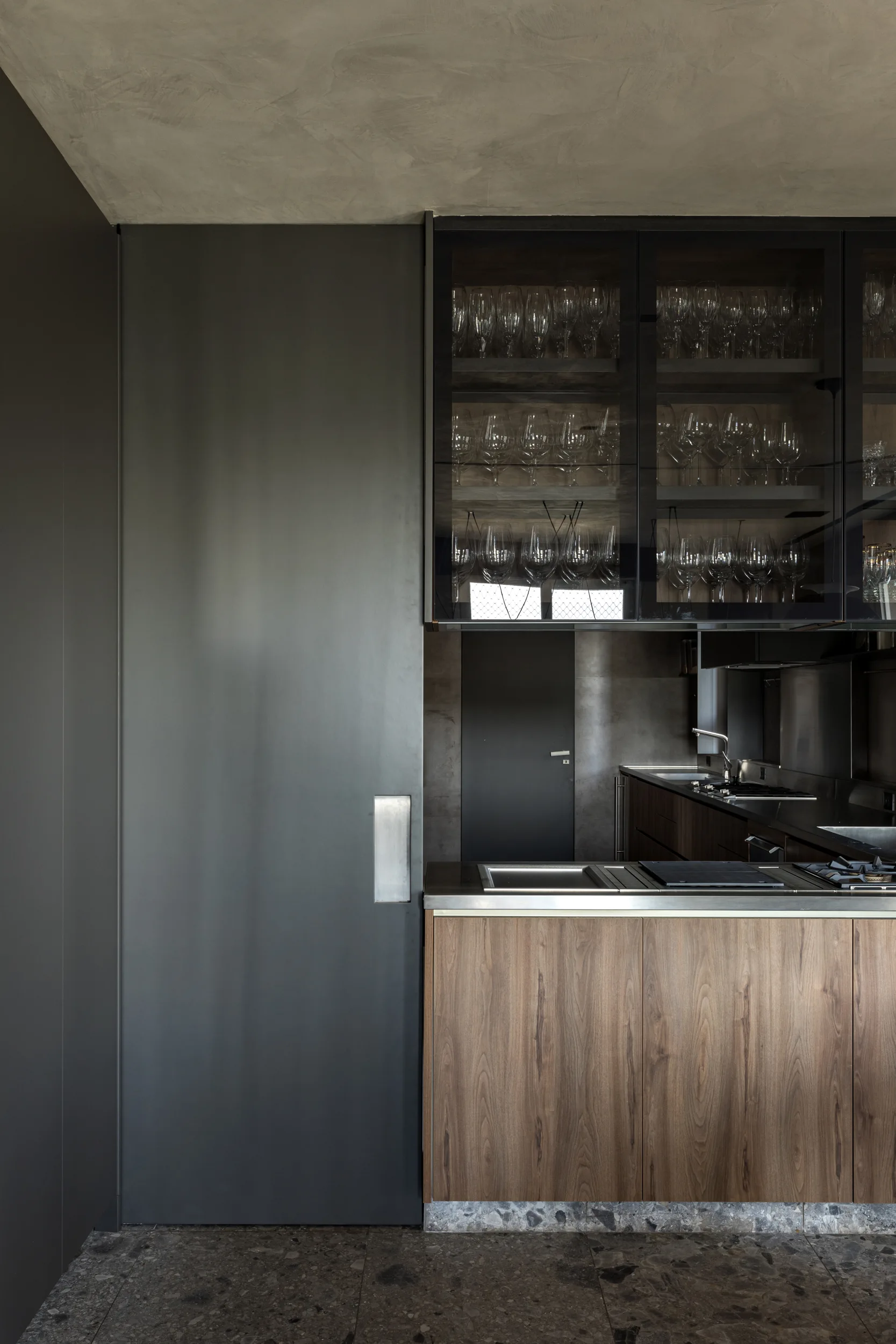 A modern kitchen with dark charcoal wall panels, wood-toned lower cabinets, and glass-front upper cabinets filled with stemware.