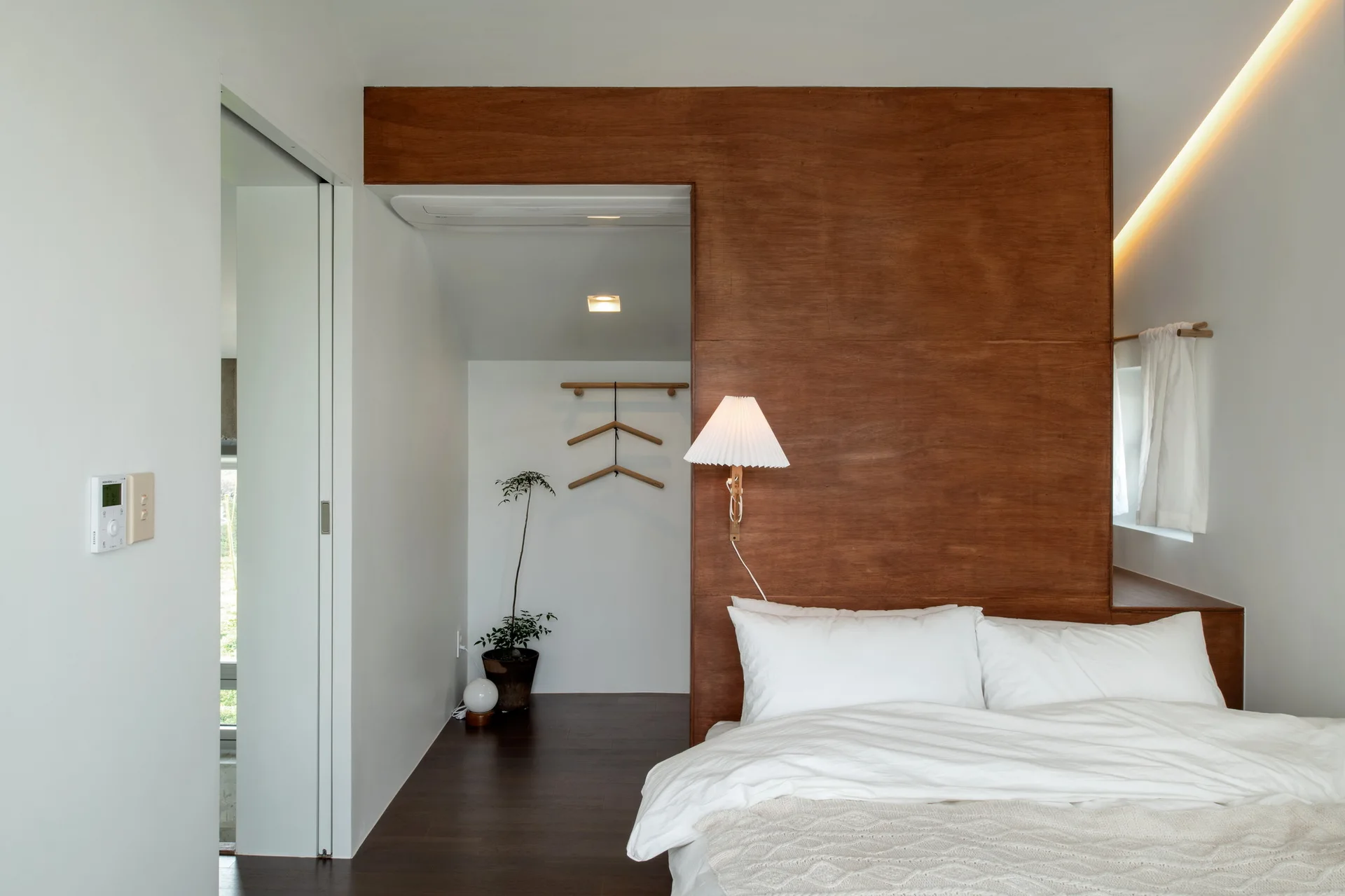 A minimalist bedroom features a large dark wood panel wall acting as a headboard behind a bed with white bedding, paired with a pleated wall sconce.