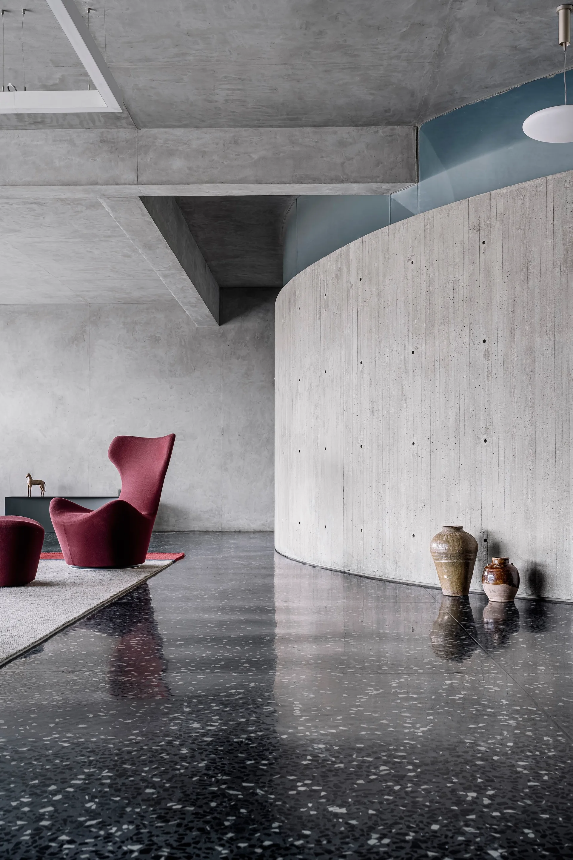 A minimalist room featuring a curved concrete wall, a polished dark terrazzo floor reflecting light, and a sculptural red armchair on a gray rug.