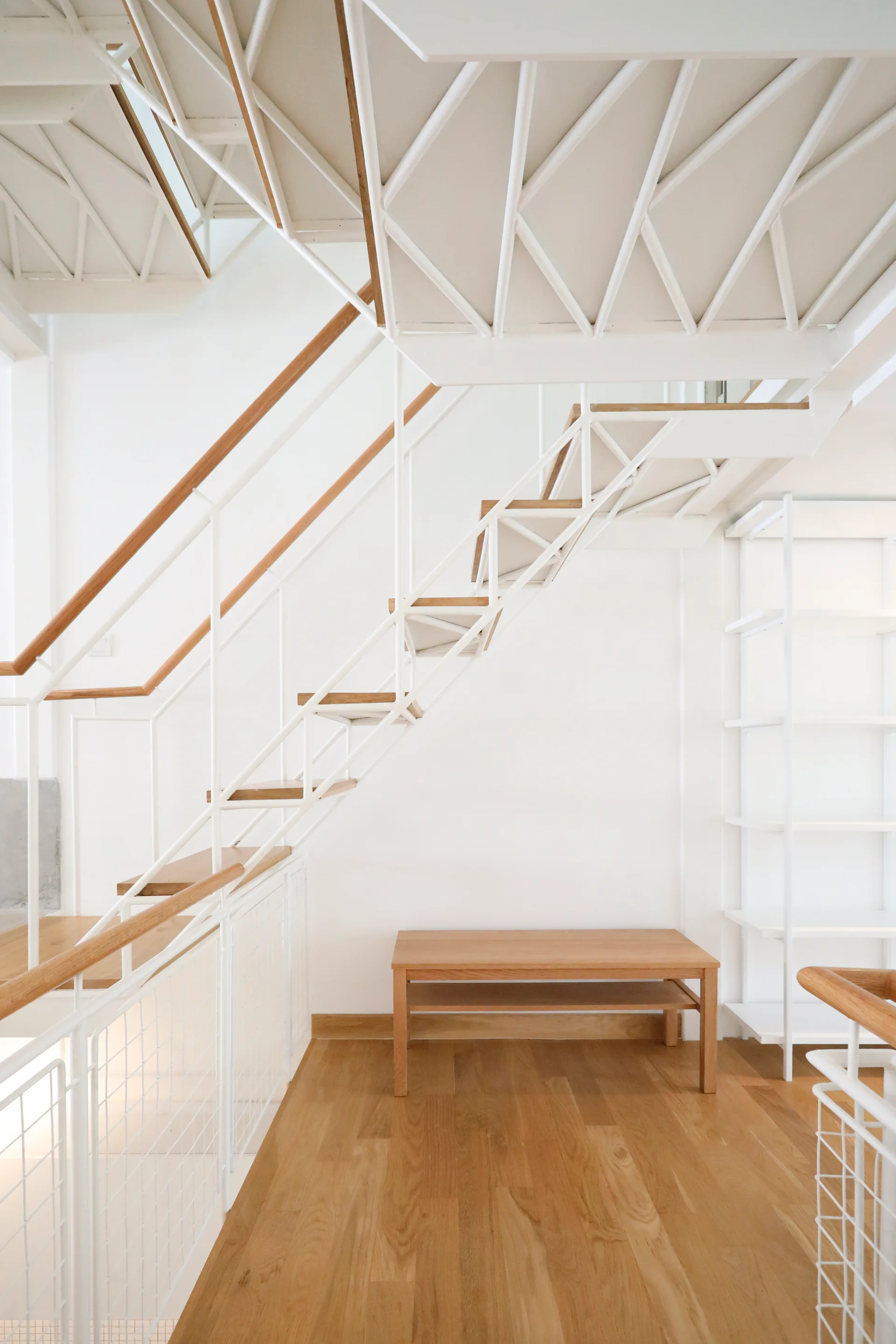 A minimalist open staircase with white metal truss framing and light wood treads stands next to a wooden bench and white shelving unit.