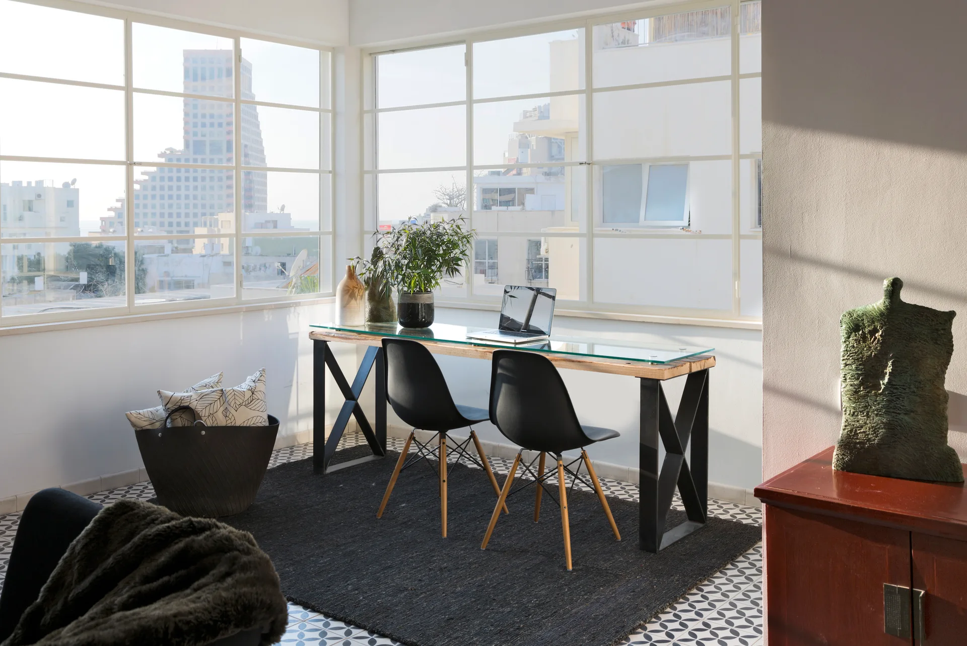 A home office setup featuring a glass-topped desk with black chairs placed on a dark rug over geometric tiled flooring in front of large windows.