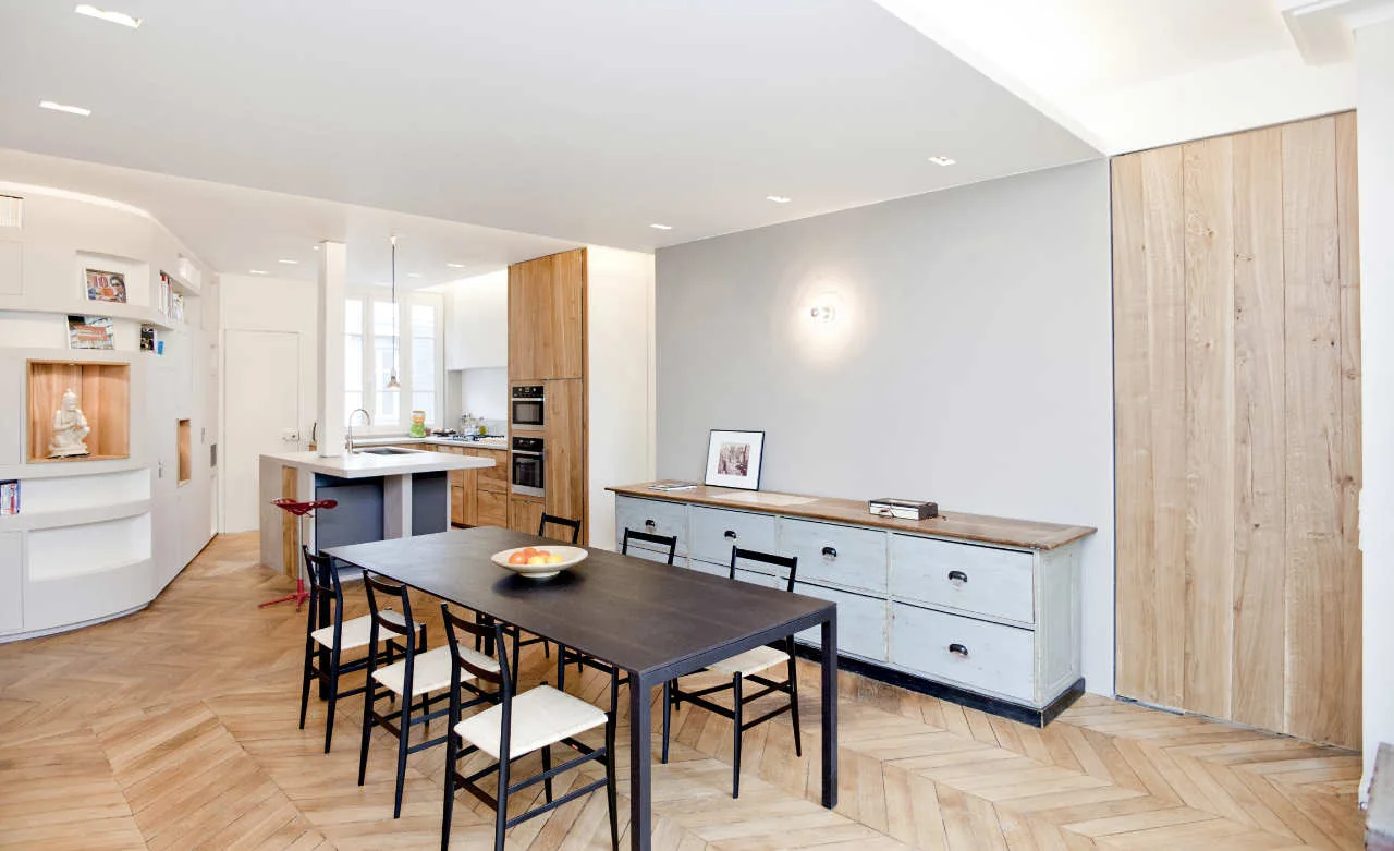A modern dining area features a dark wood table and chairs on herringbone flooring beside a grey cabinet with drawers.