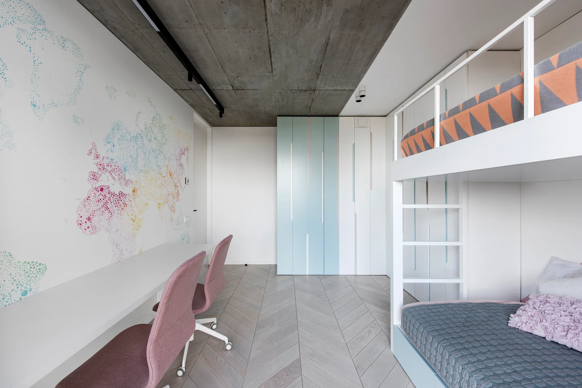 A modern children's bedroom features a white bunk bed, two pink chairs at a long white desk, a world map wall mural, and an exposed concrete ceiling.