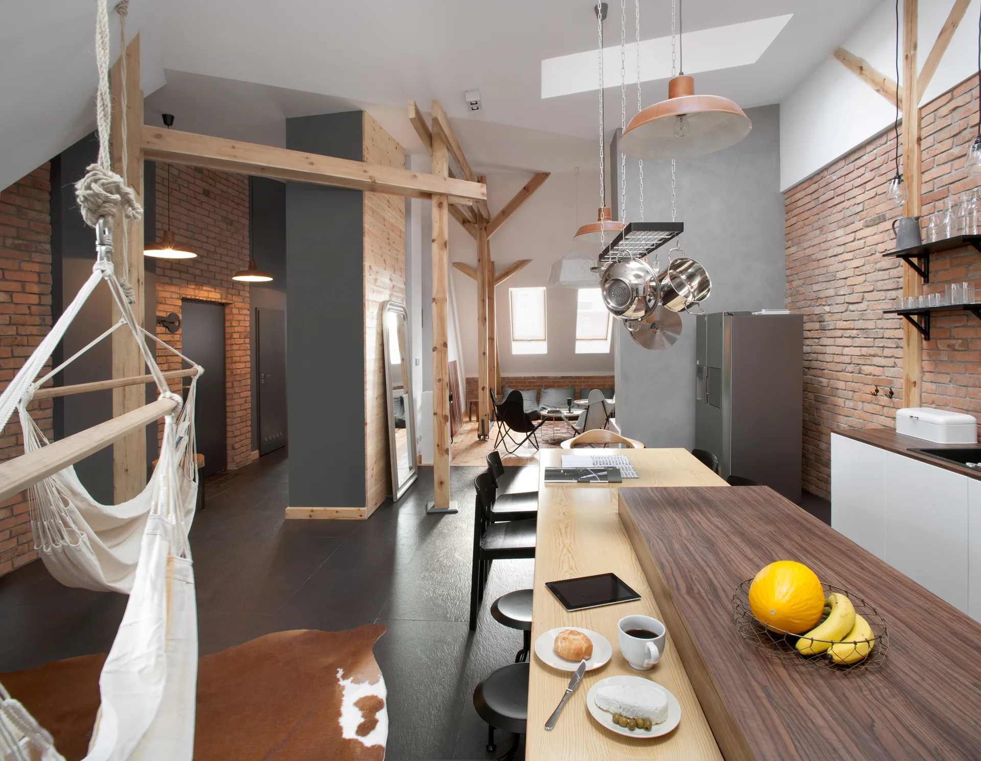 An industrial loft kitchen features a long dual-toned wooden island, exposed brick walls, structural timber framing, and a hanging white hammock.