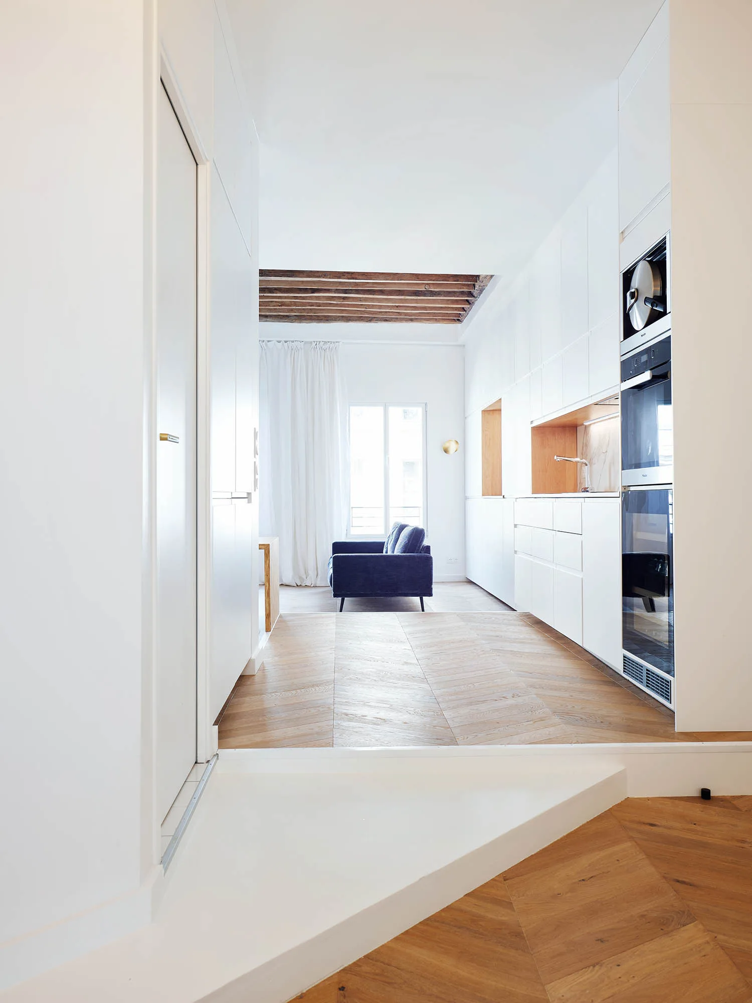 A minimalist white kitchen with handleless cabinetry, herringbone wood flooring, built-in ovens, and exposed wooden ceiling beams in the living area beyond.