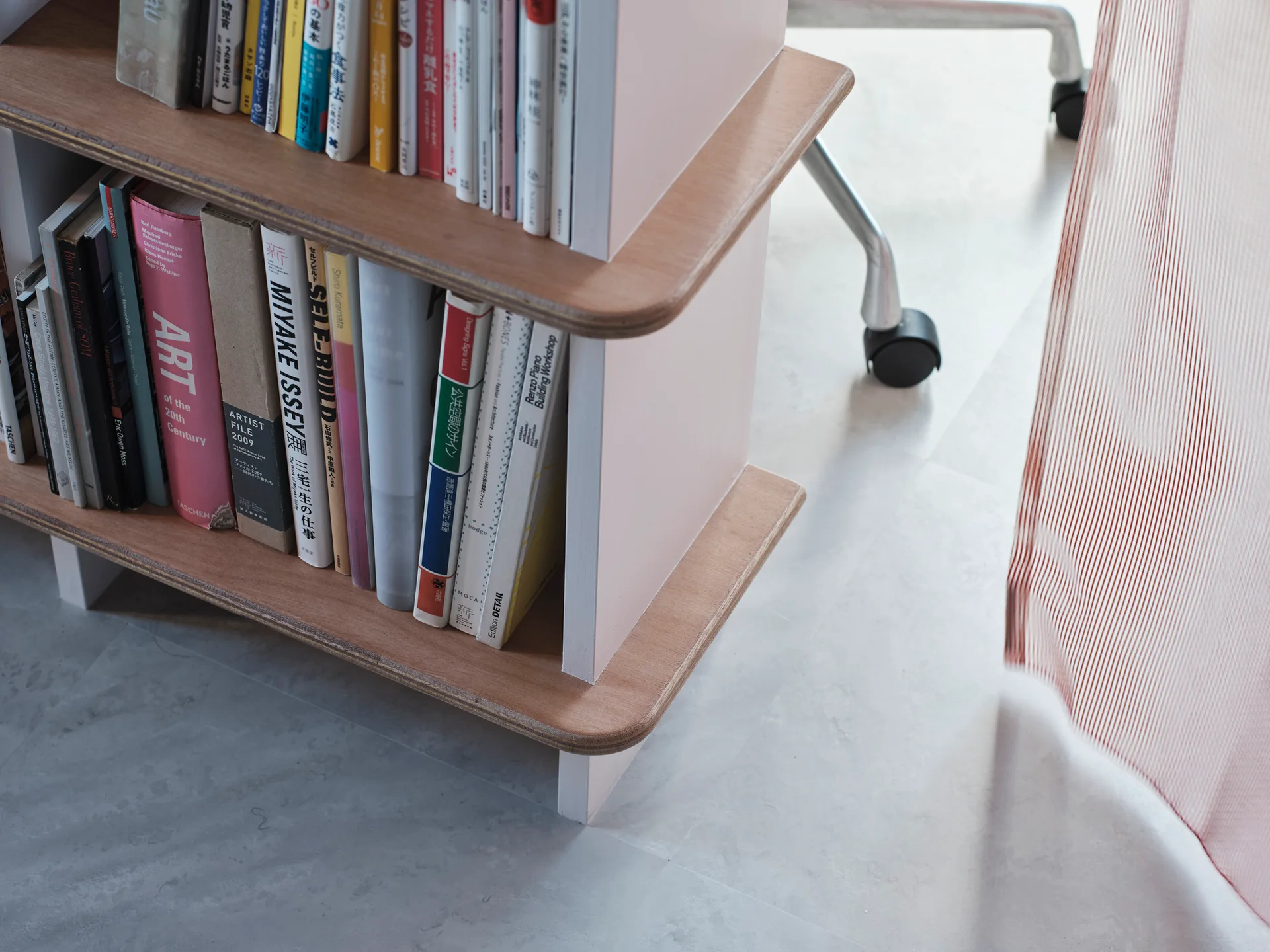 A close-up of a custom light pink and wood plywood bookshelf with rounded corners next to an office chair on a grey floor.
