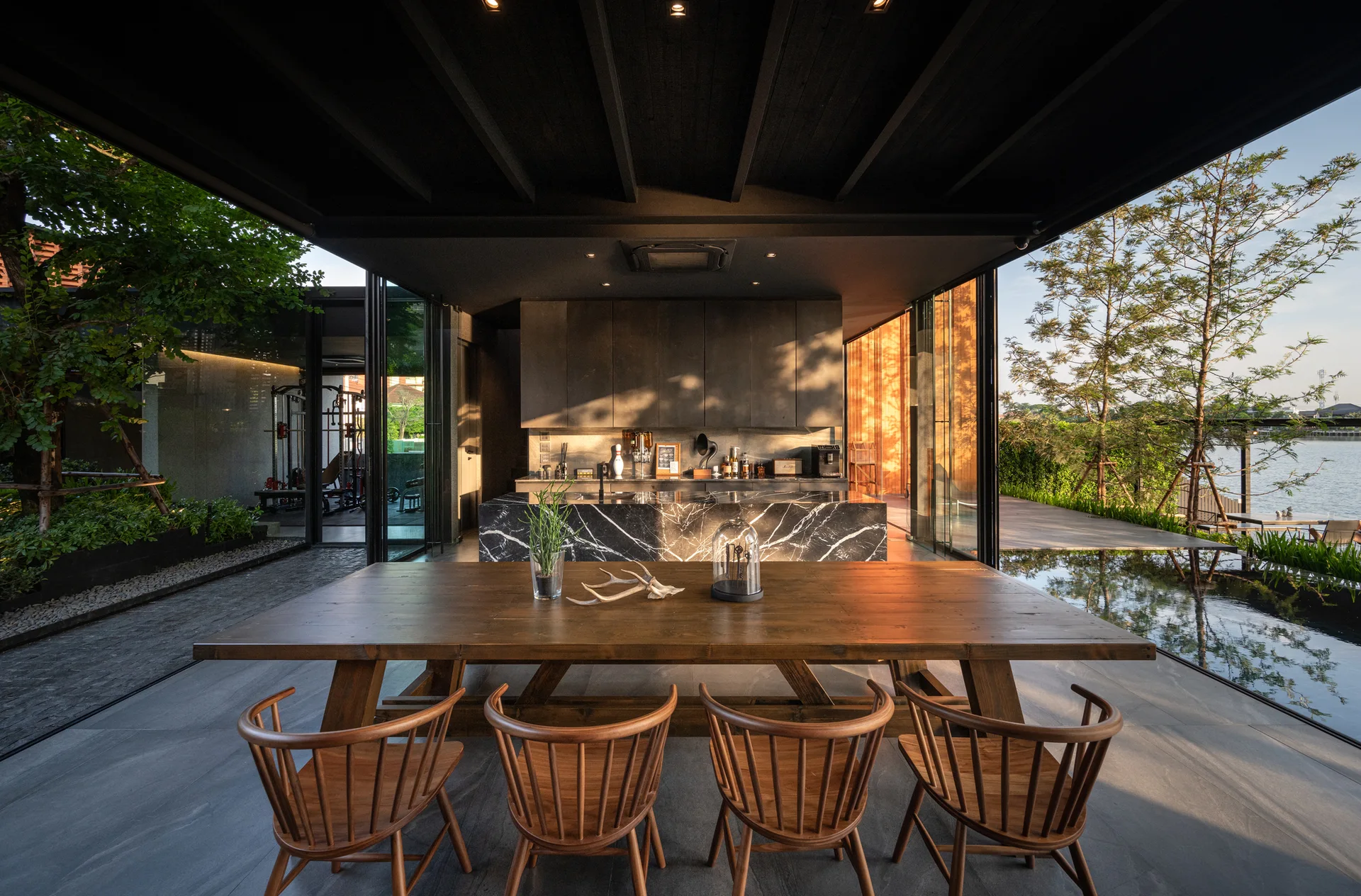 A modern dining space features a wooden table with spindle chairs beneath a dark beamed ceiling next to a black marbled kitchen island.