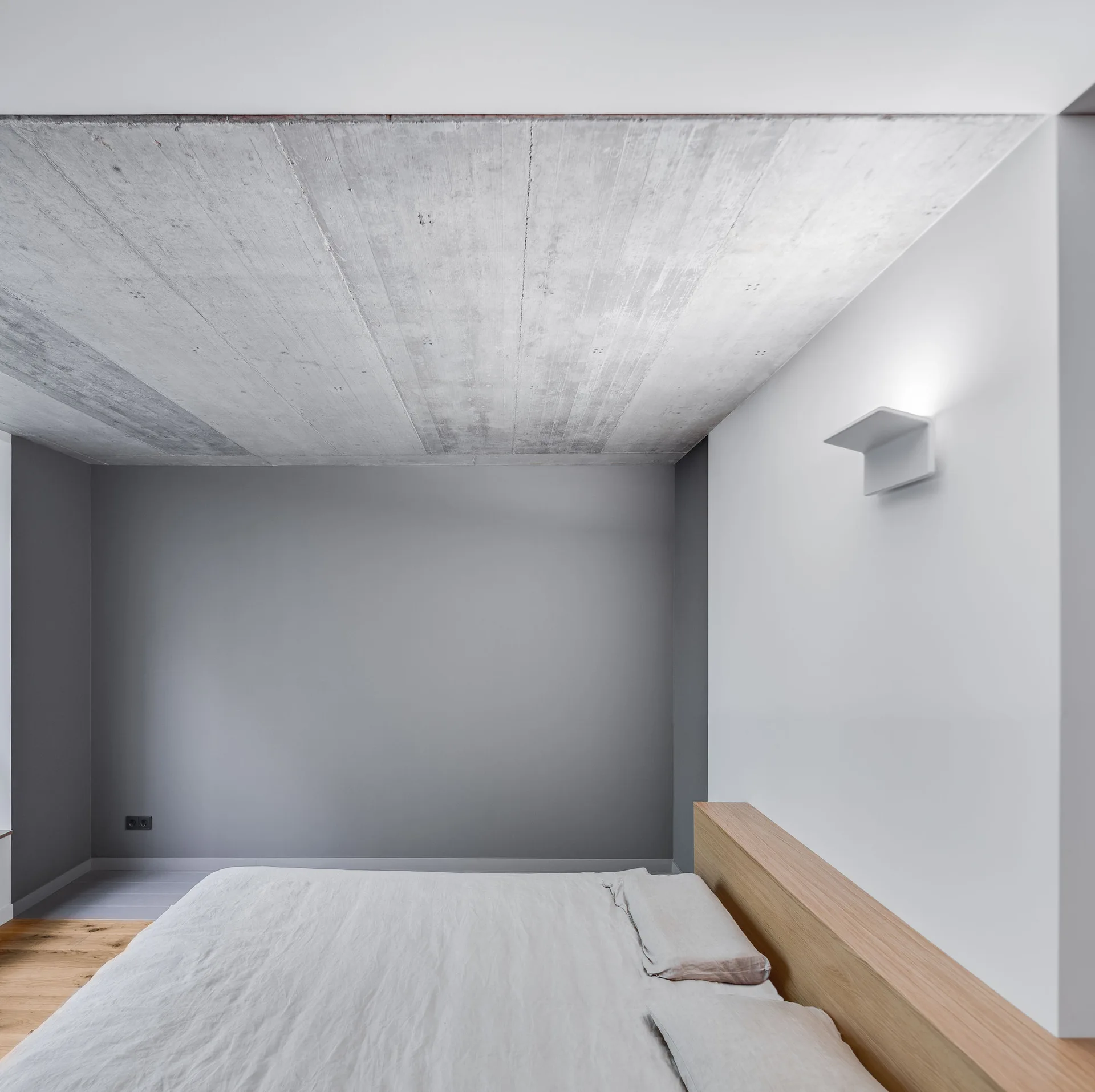 A minimalist bedroom features an exposed concrete ceiling, a light wood headboard, a grey wall, and a modern white wall sconce.