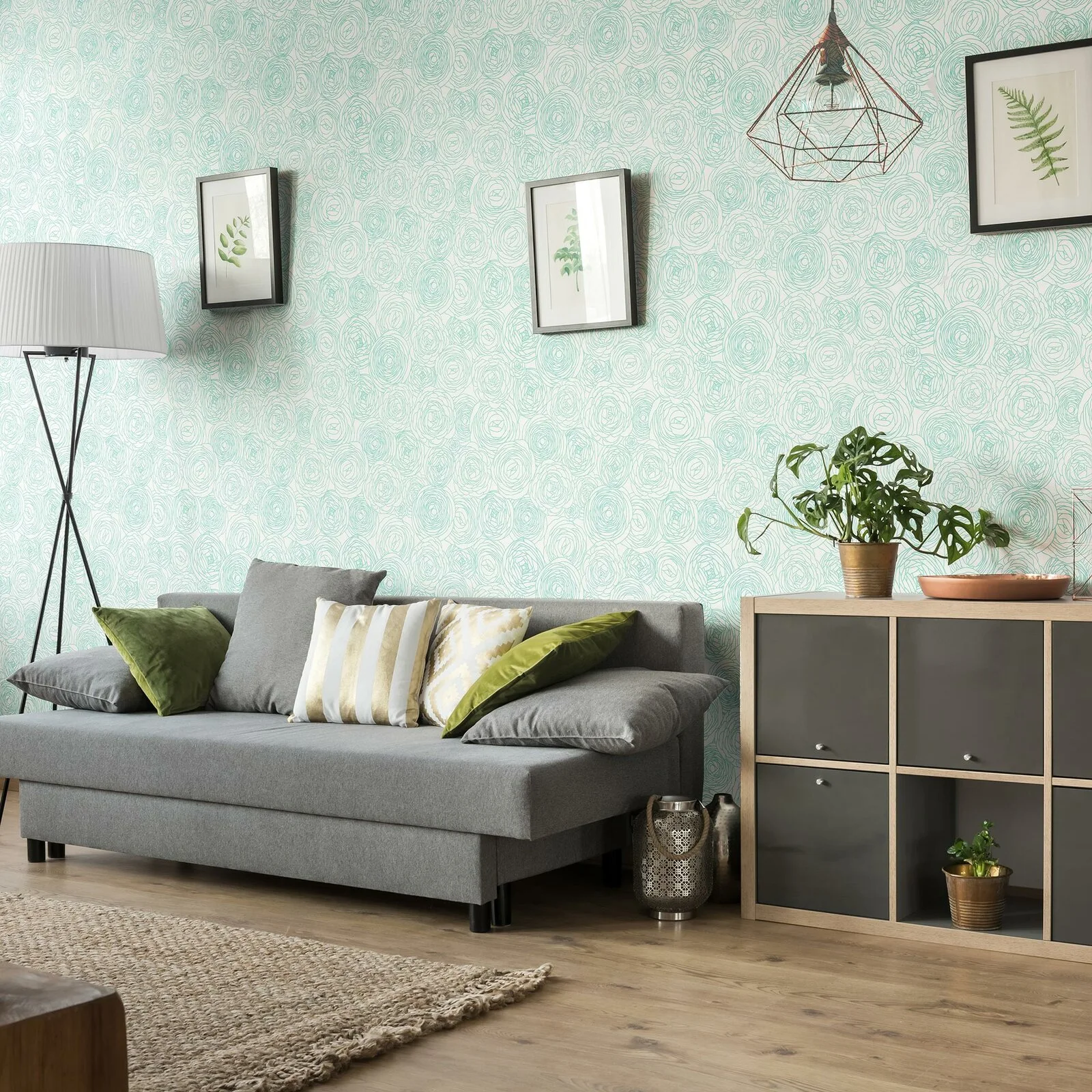 A grey sofa with green and gold throw pillows stands against a mint green patterned wallpaper wall beside a wooden storage unit.
