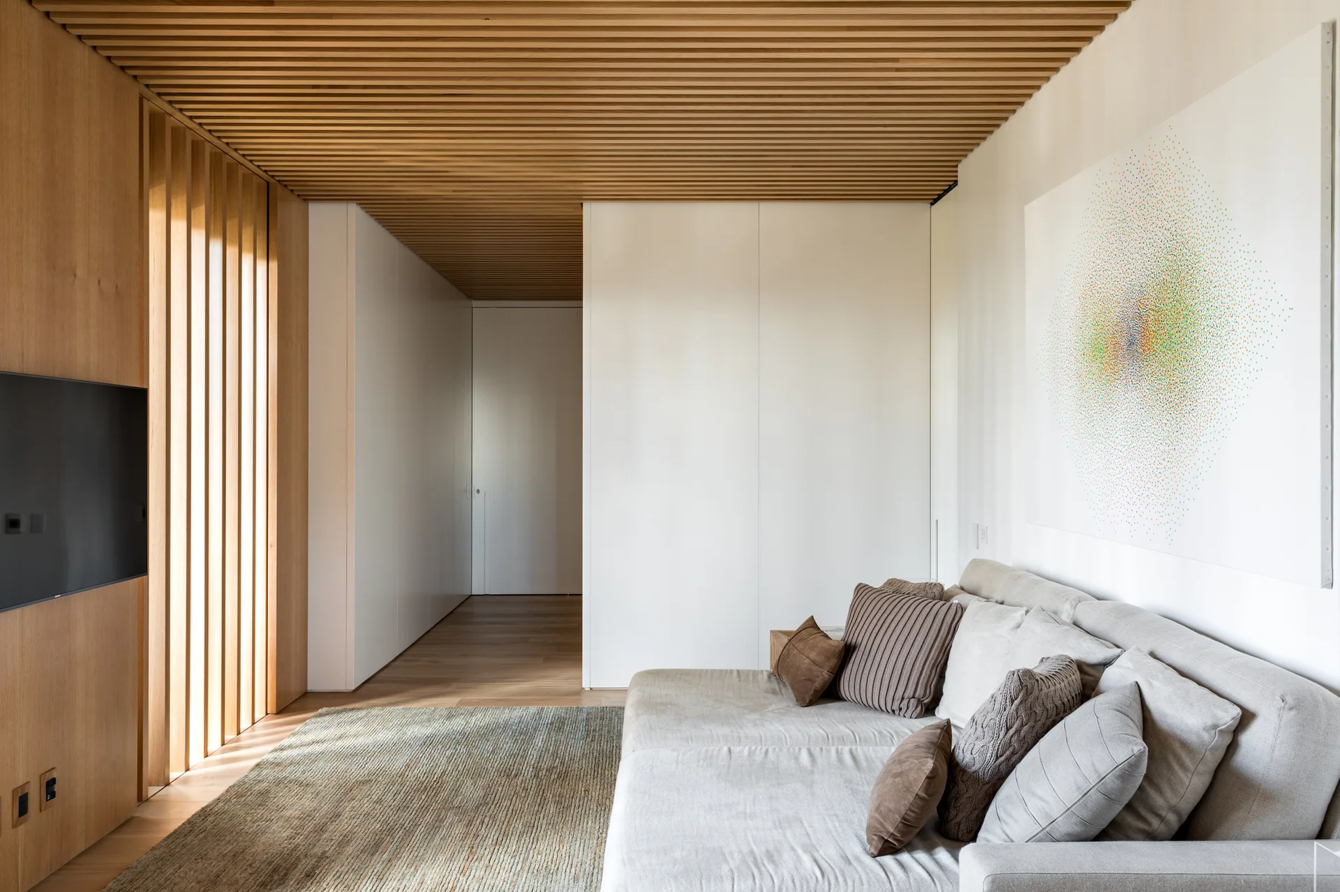 A modern lounge features a light gray sofa with textured brown pillows beneath a slatted wood ceiling, next to flush white storage cabinets.