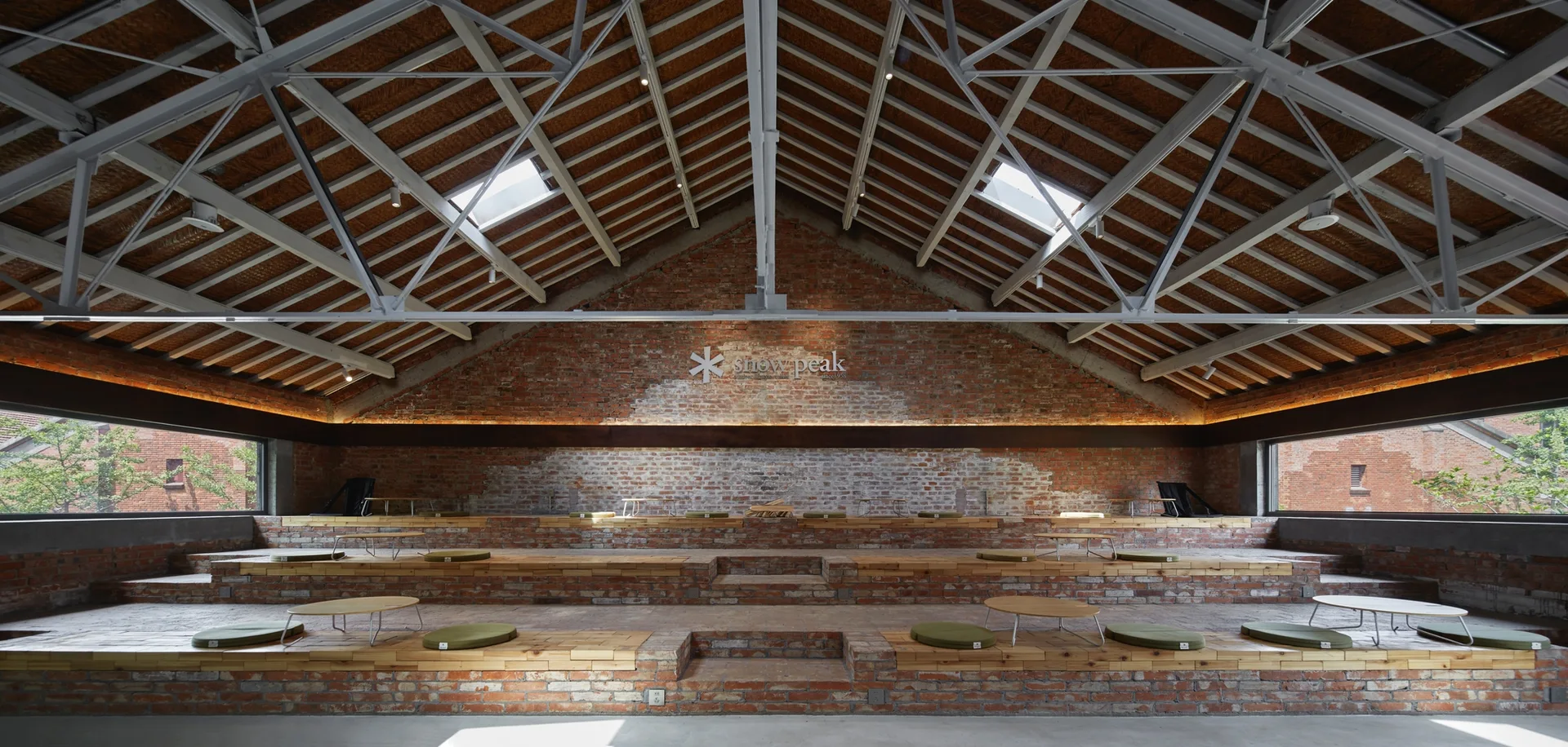 Tiered brick seating platforms with wood tops and green cushions inside an industrial hall with exposed brick walls and a metal truss ceiling.
