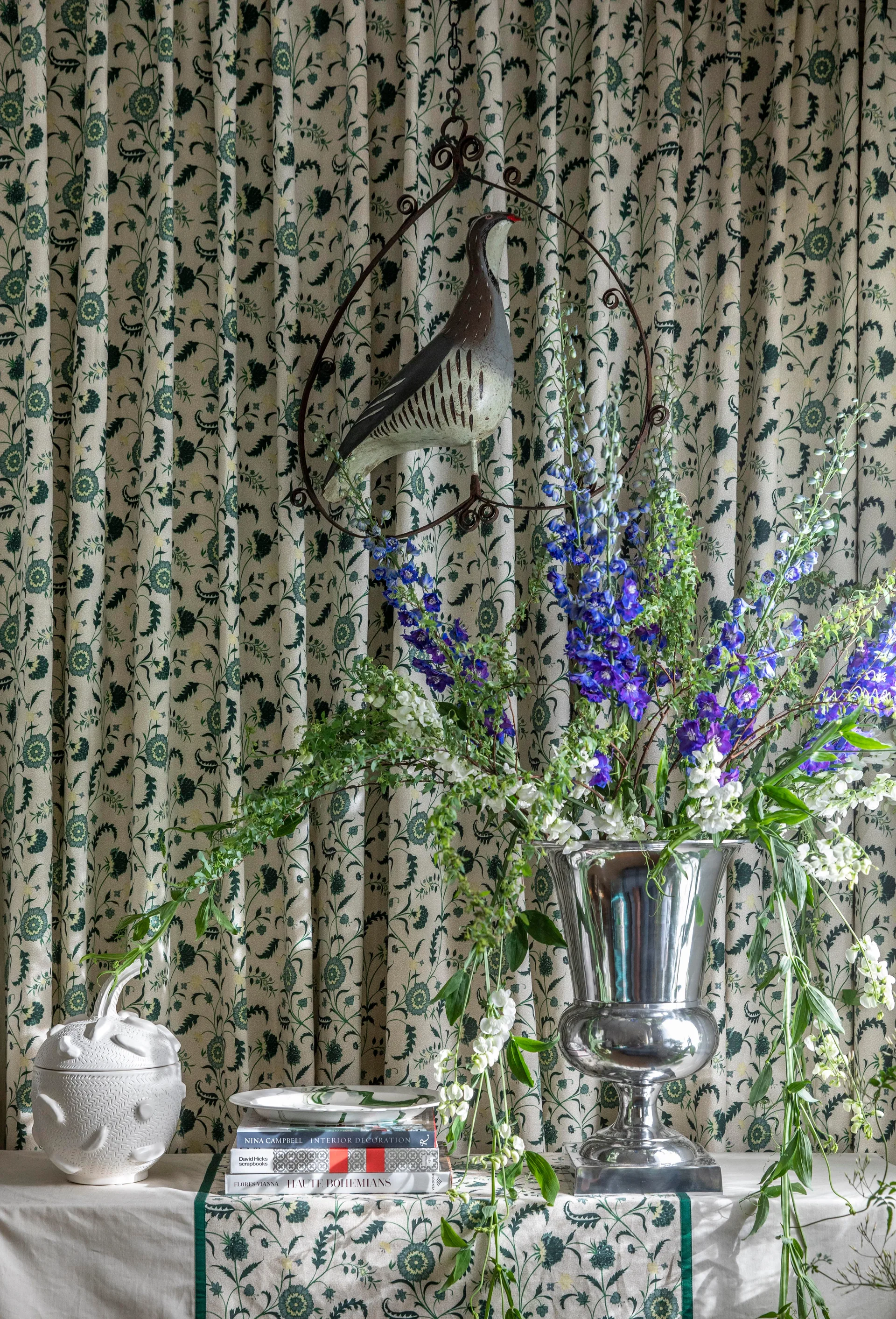 A tabletop vignette features a silver urn with blue flowers, textured ceramics, and books set against green and cream botanical patterned drapery.