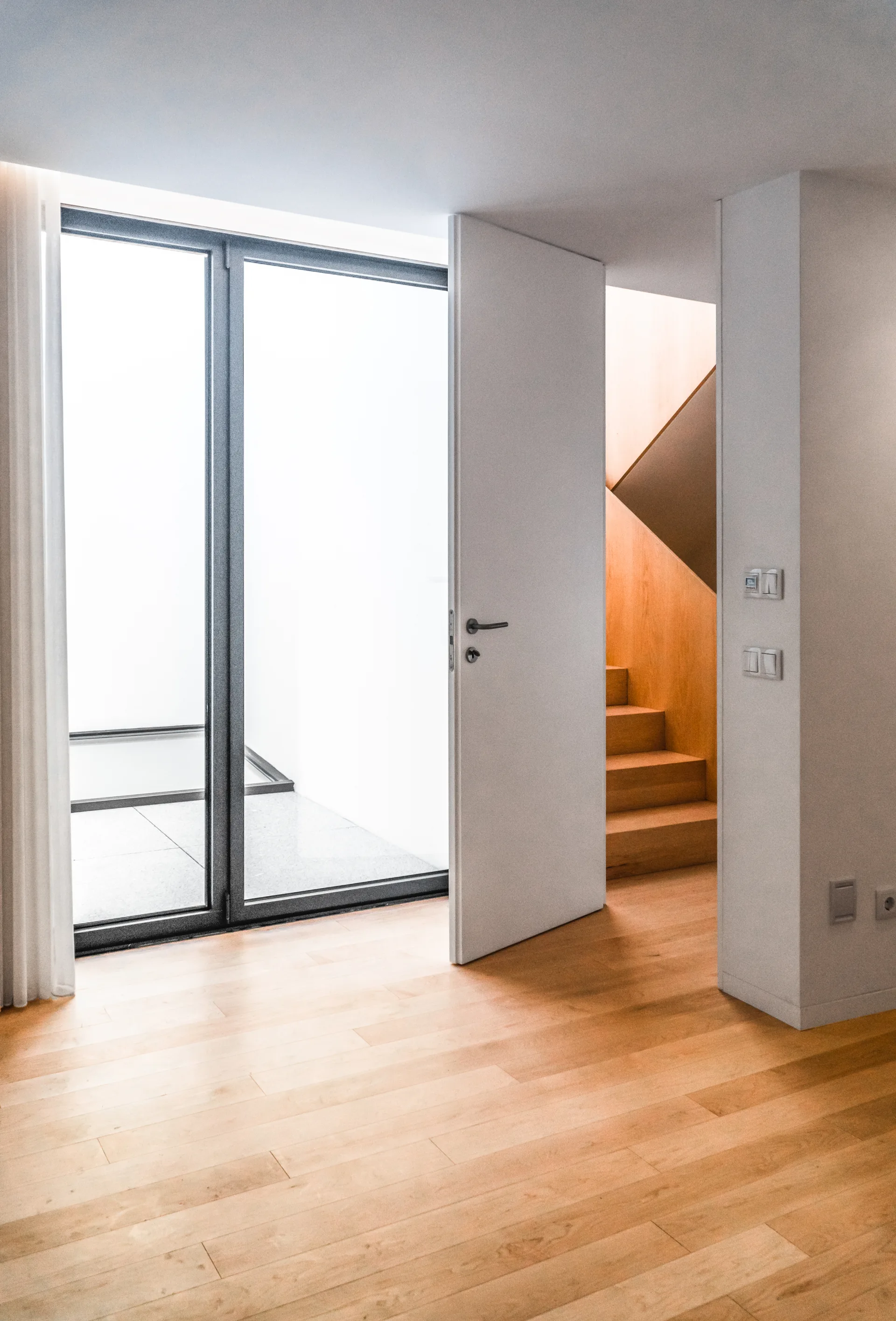 A white door swings open in a bright hallway with light wood flooring, revealing matching wooden stairs and a large window.