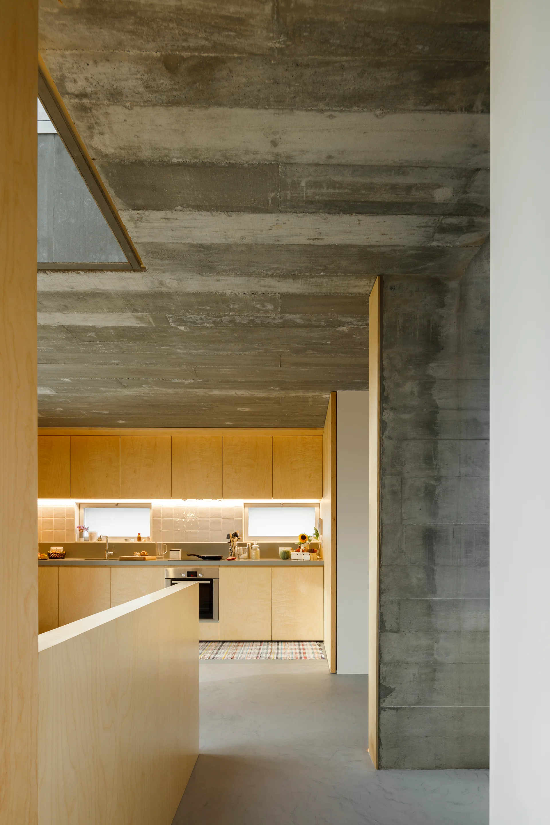 A modern kitchen features light wood cabinets, small horizontal windows, and a rough board-formed concrete ceiling and structural pillar.