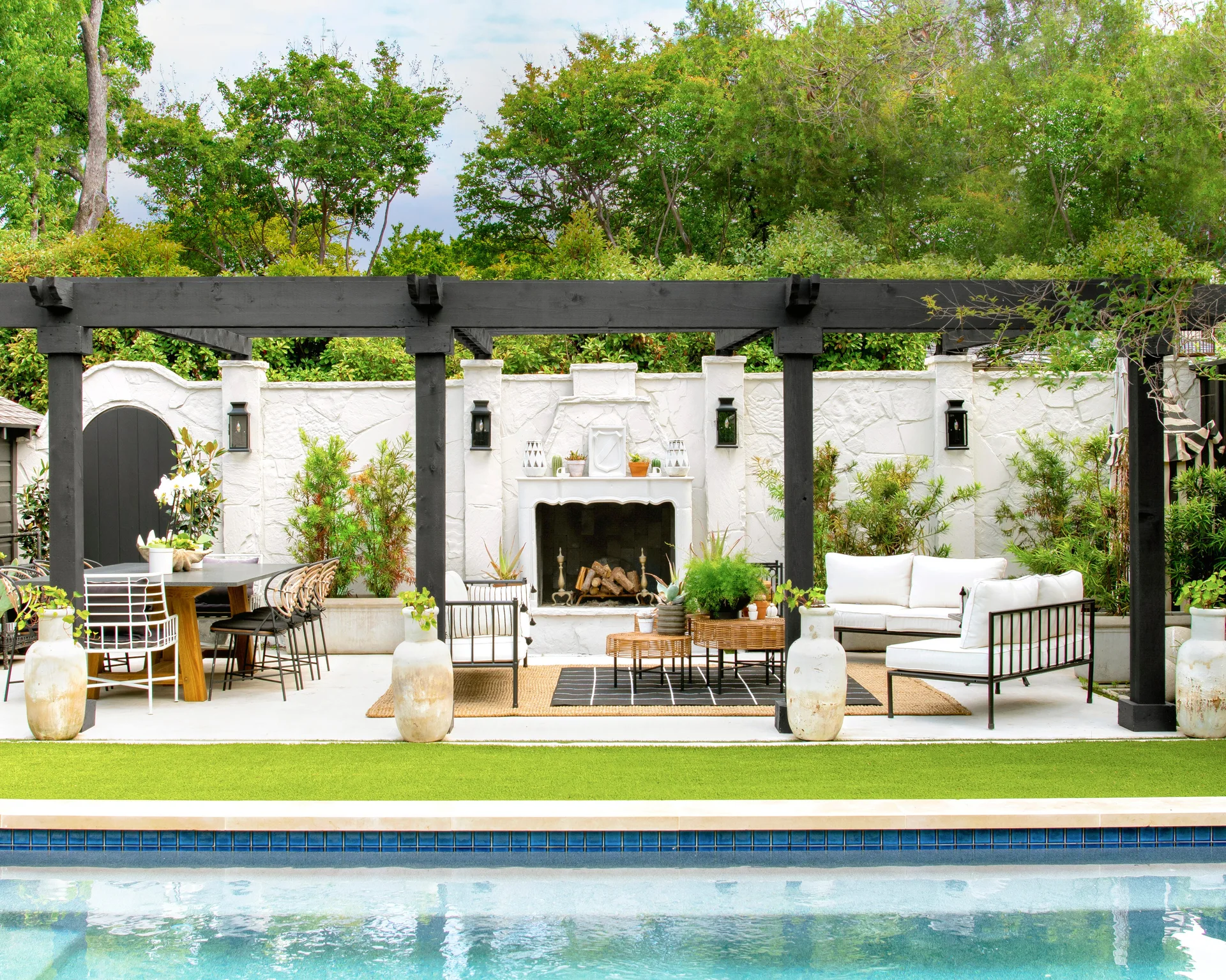 A poolside concrete patio features a black wooden pergola, a central white stone fireplace, outdoor seating, and a dining table.
