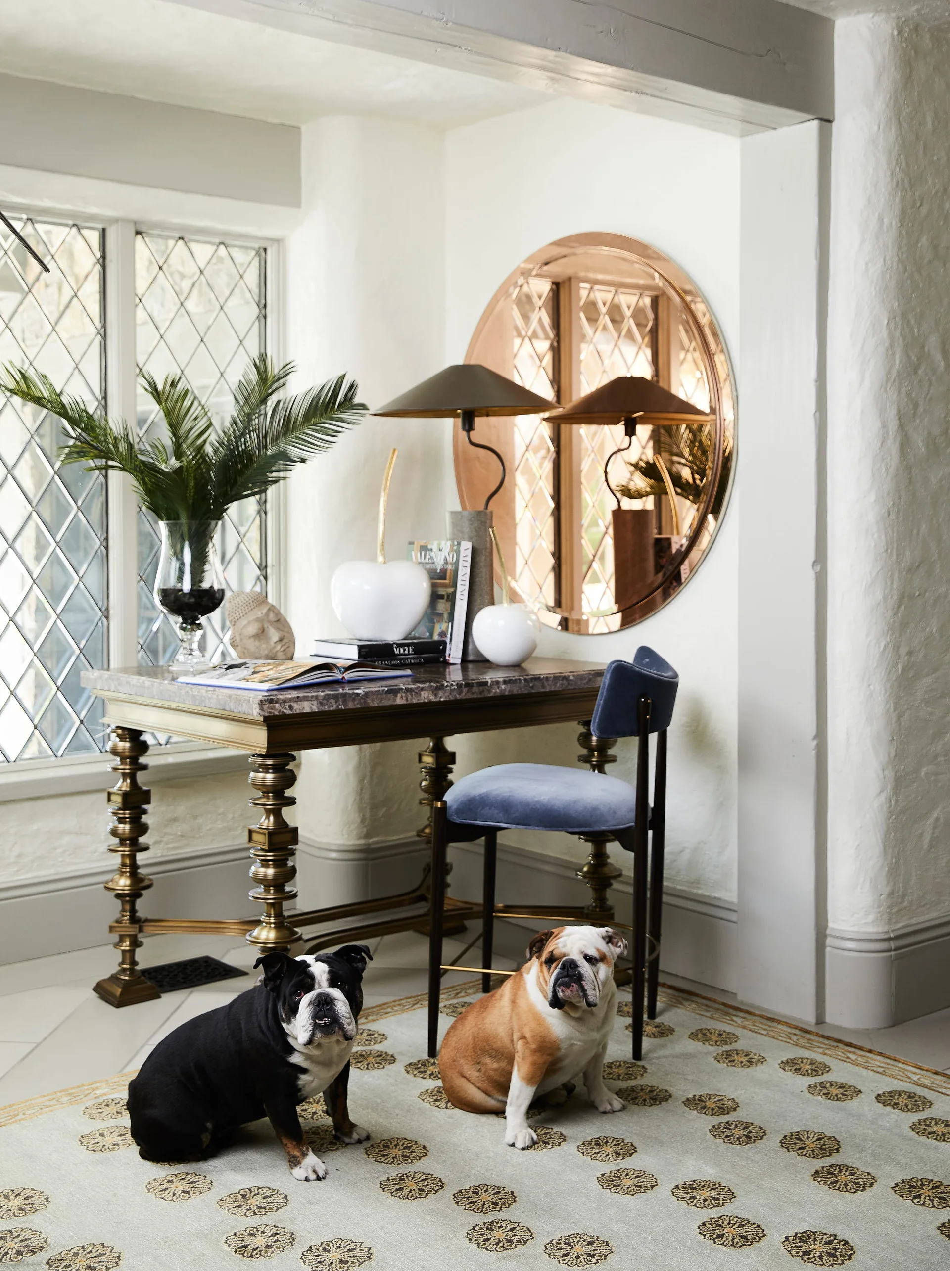 A home office nook features a marble-top bronze desk, a blue velvet chair, and a round mirror next to a diamond-patterned leaded glass window.