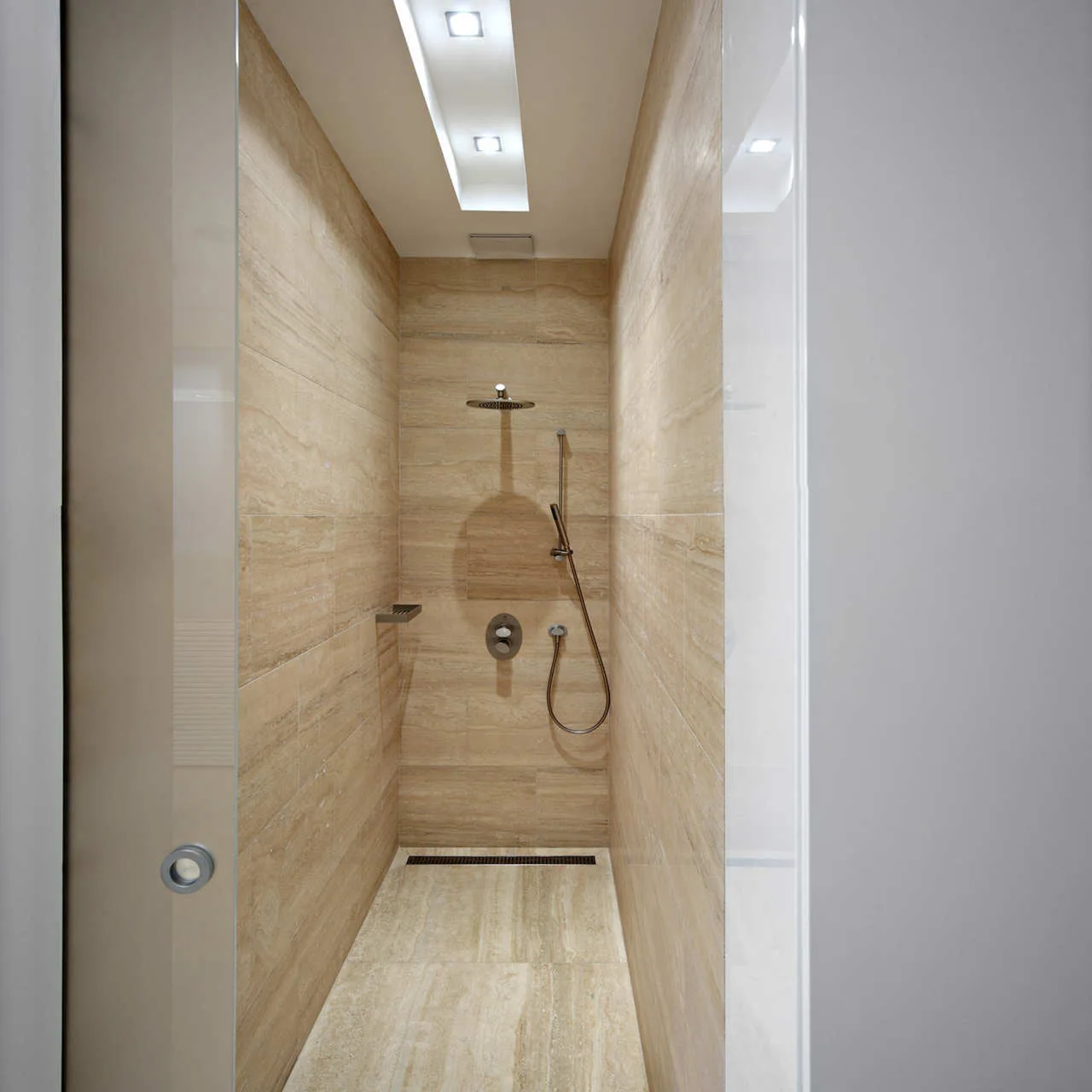 A narrow modern shower stall clad in beige stone tiles with a rainfall shower head, handheld wand, and linear drain.