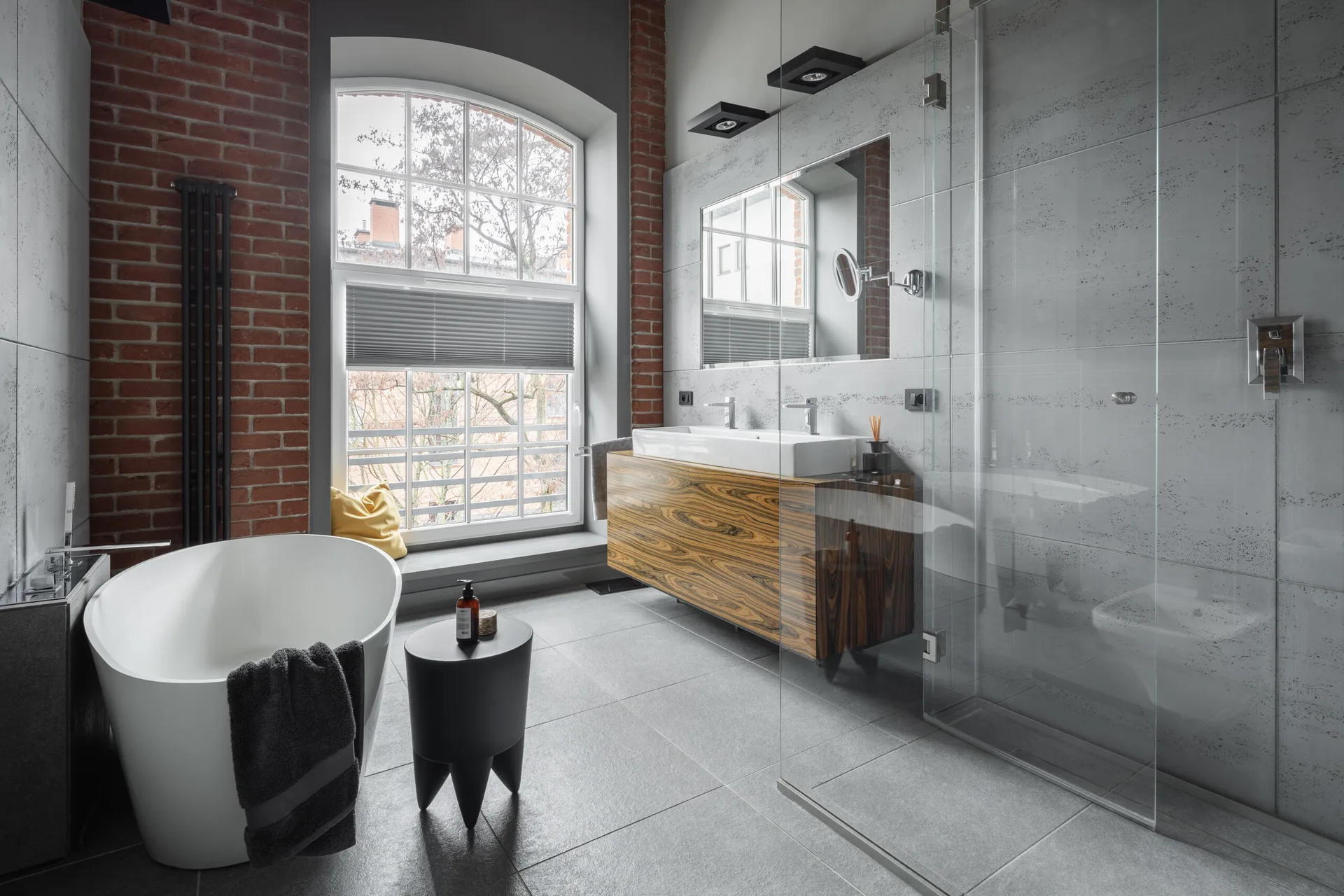 A modern bathroom with a freestanding white tub, a wall-mounted wood vanity, gray floor tiles, and exposed red brick pillars framing a large arched window.