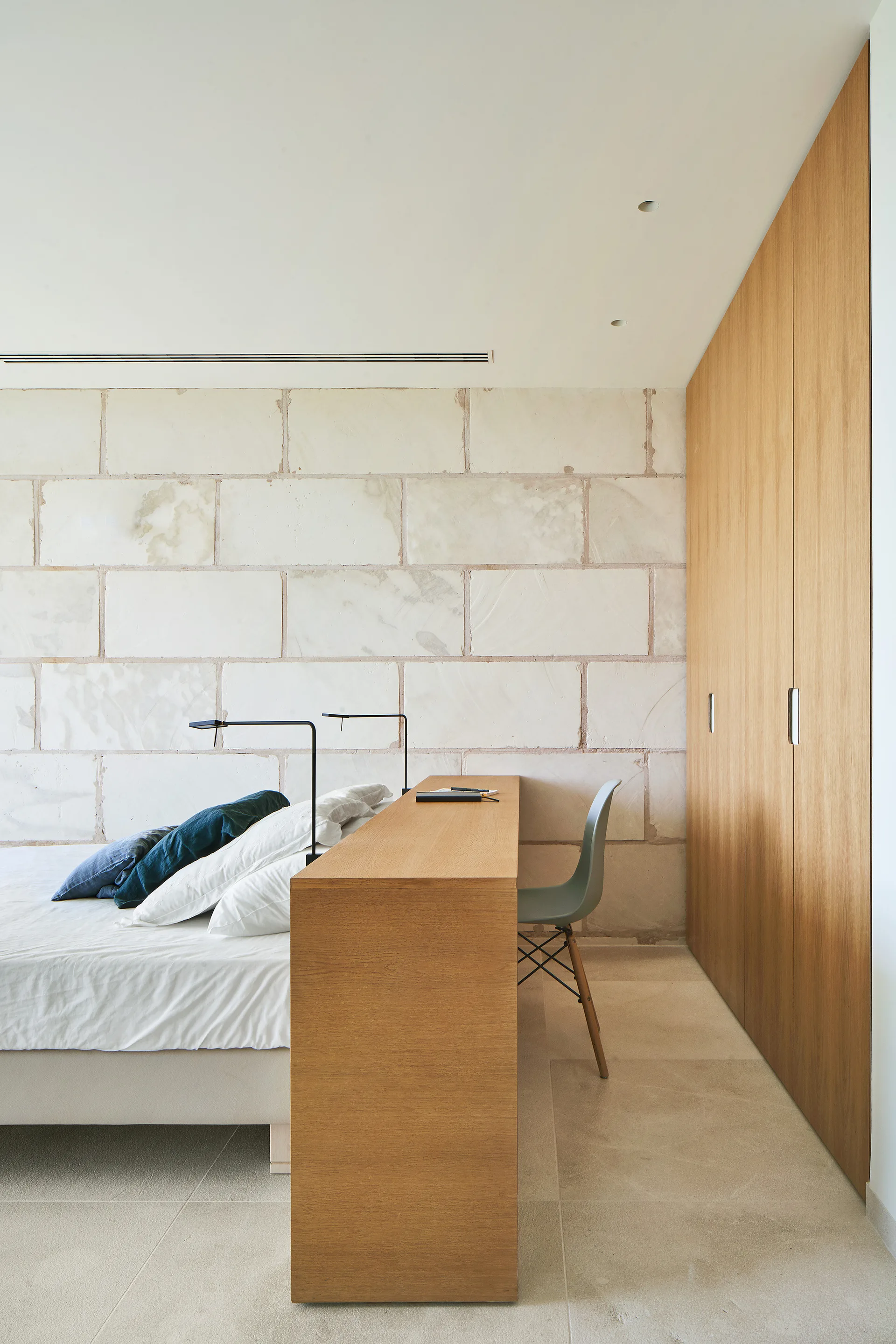 A minimalist bedroom featuring a bed against a textured stone block wall, an integrated wood desk, and a built-in wardrobe.