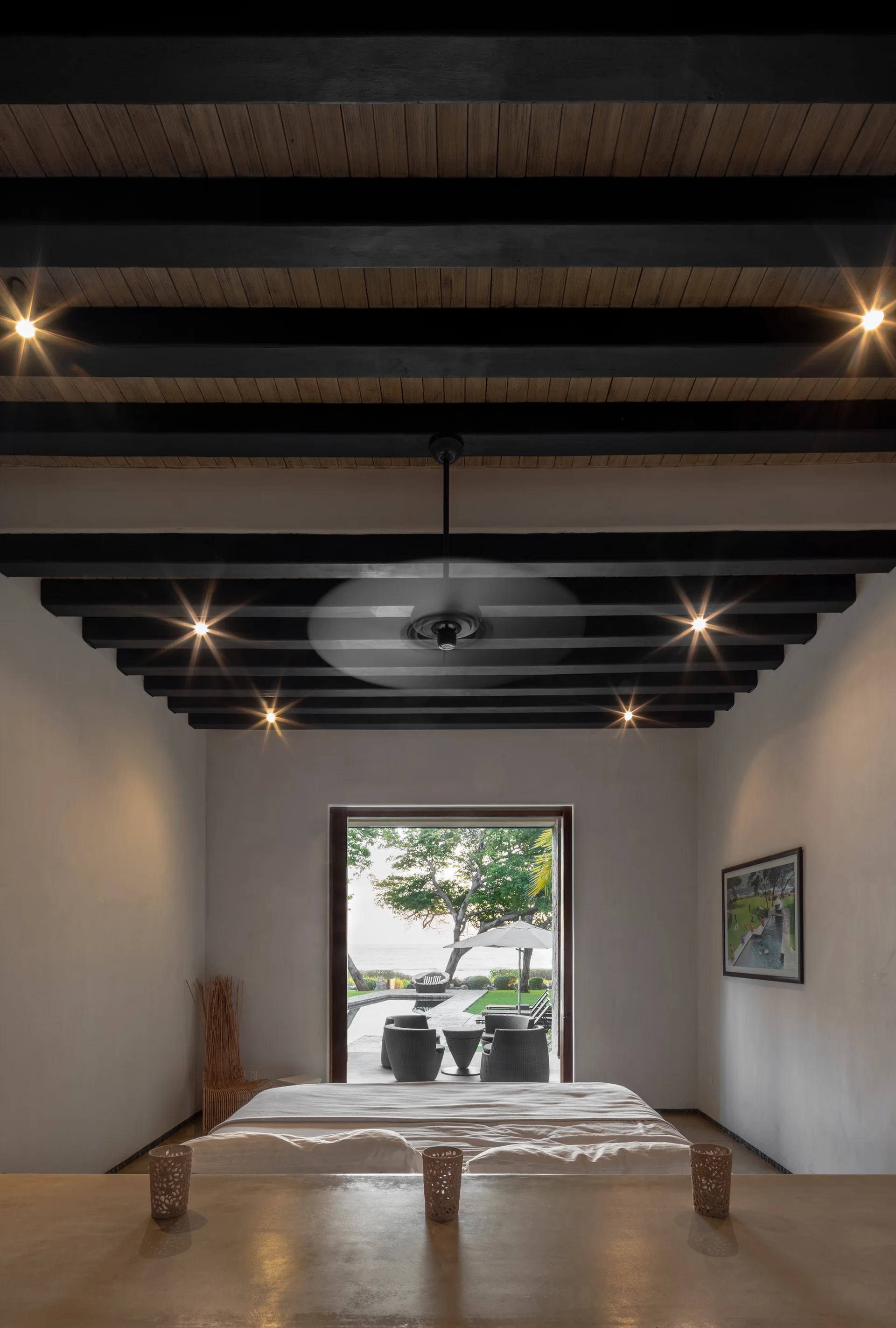 A minimalist bedroom features dark wooden ceiling beams, smooth plaster walls, and an open doorway looking out toward a patio pool area.