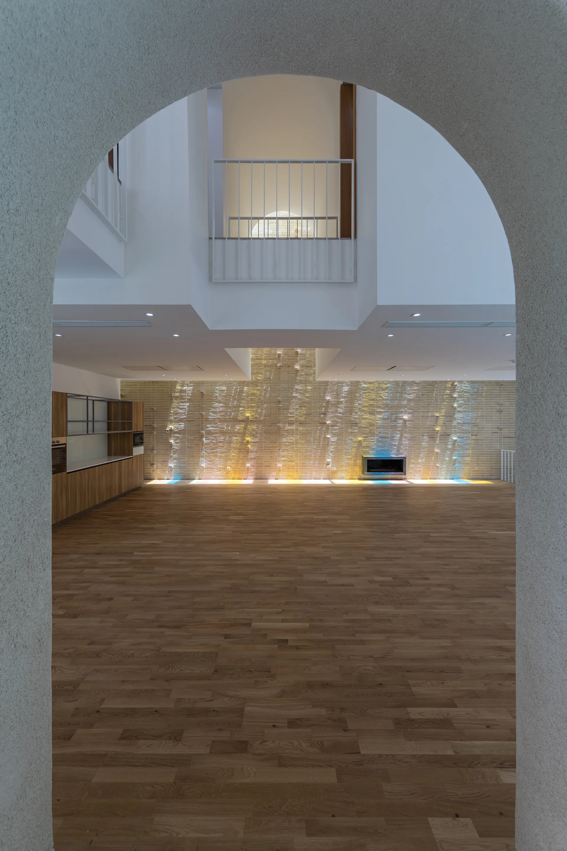 An arched opening frames a minimalist interior featuring wood flooring, an illuminated textured wall, and an upper-level mezzanine balcony.