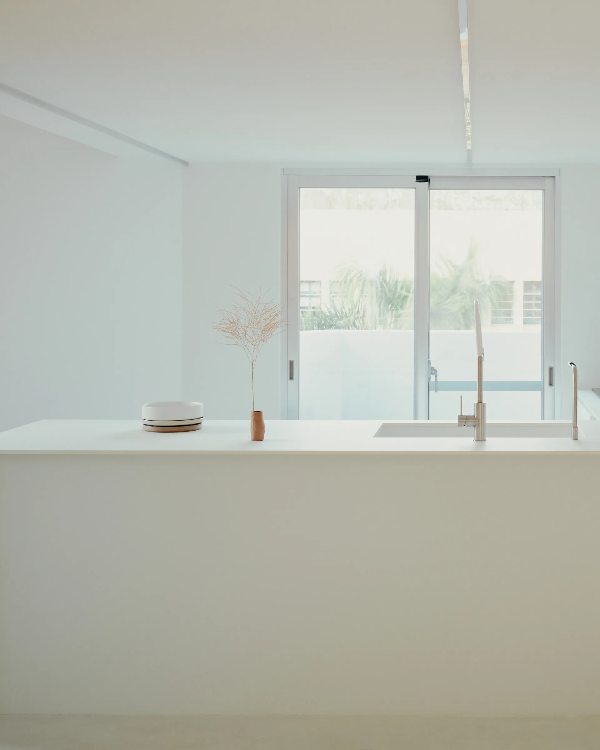 A minimalist white kitchen island with an integrated sink and metal faucet stands before a large sliding glass door.