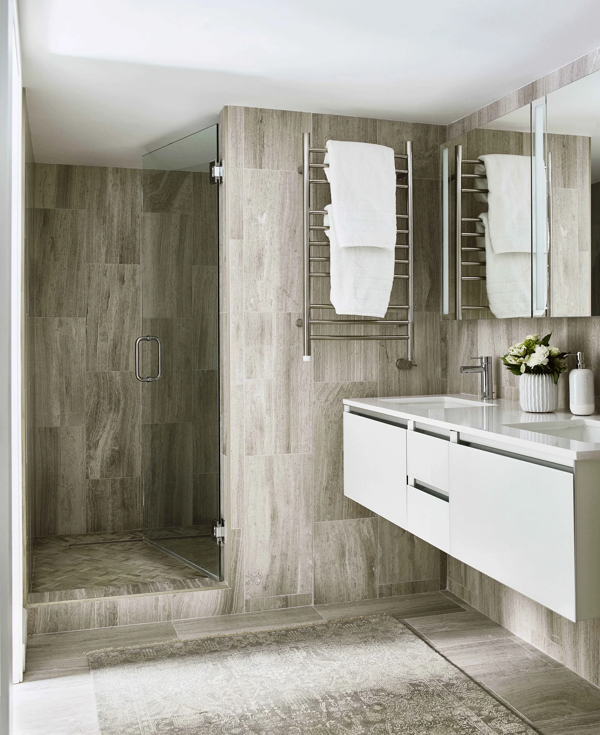 A modern bathroom features wood-look wall and floor tiles, a floating white vanity with double faucets, and a glass walk-in shower.
