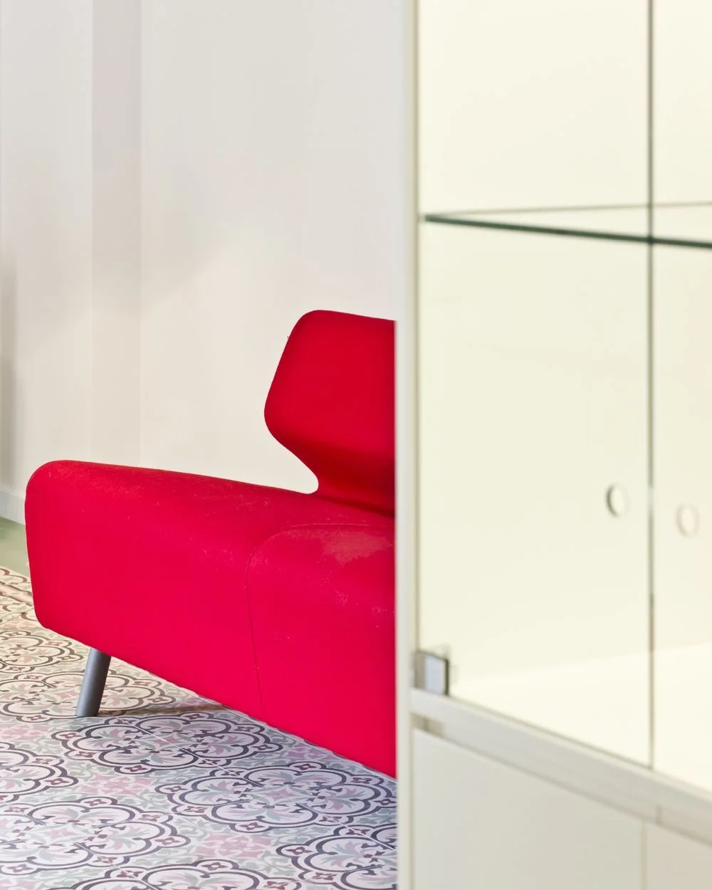 A modern red sofa sits on violet and grey patterned floor tiles next to a white glass-fronted display cabinet against a plain white wall.