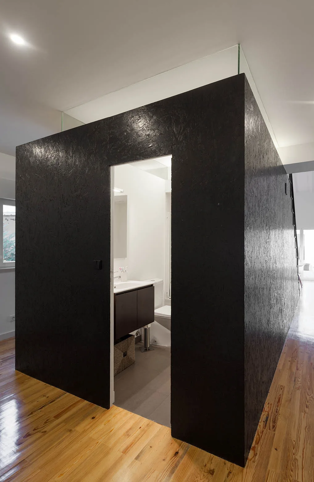 A freestanding black textured OSB cube houses a compact bathroom with a vanity and toilet, positioned on a glossy wood floor.
