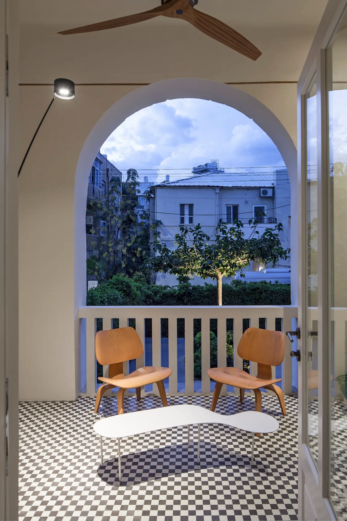A balcony with a black and white checkered floor features two wooden chairs and an organic white coffee table under a large plaster archway.