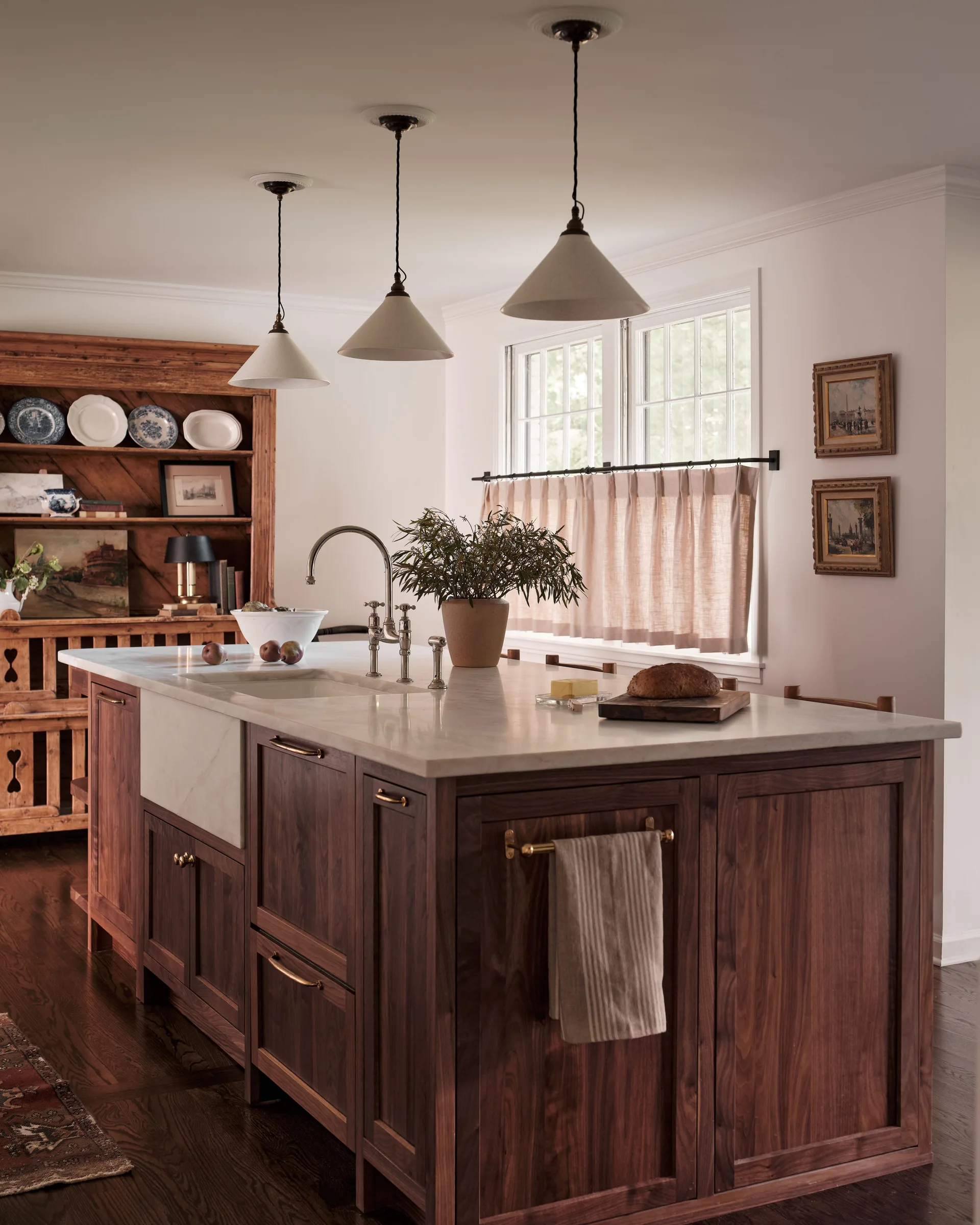A dark wood kitchen island with a white marble countertop and farmhouse sink sits under three cone pendant lights near a window with cafe curtains.