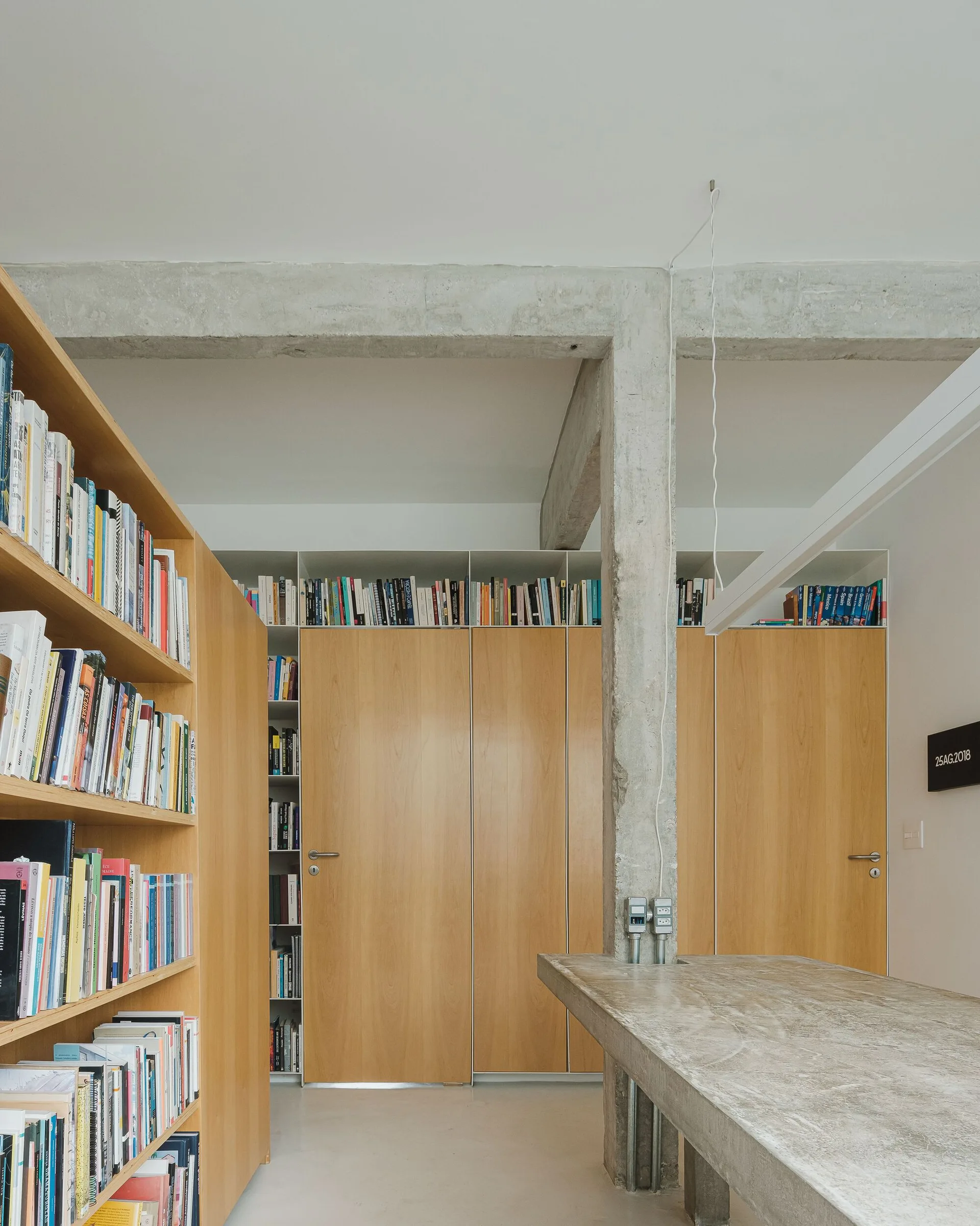 An industrial study with floor-to-ceiling wood cabinets, open bookshelves filled with books, and an exposed raw concrete column integrated with a concrete desk.