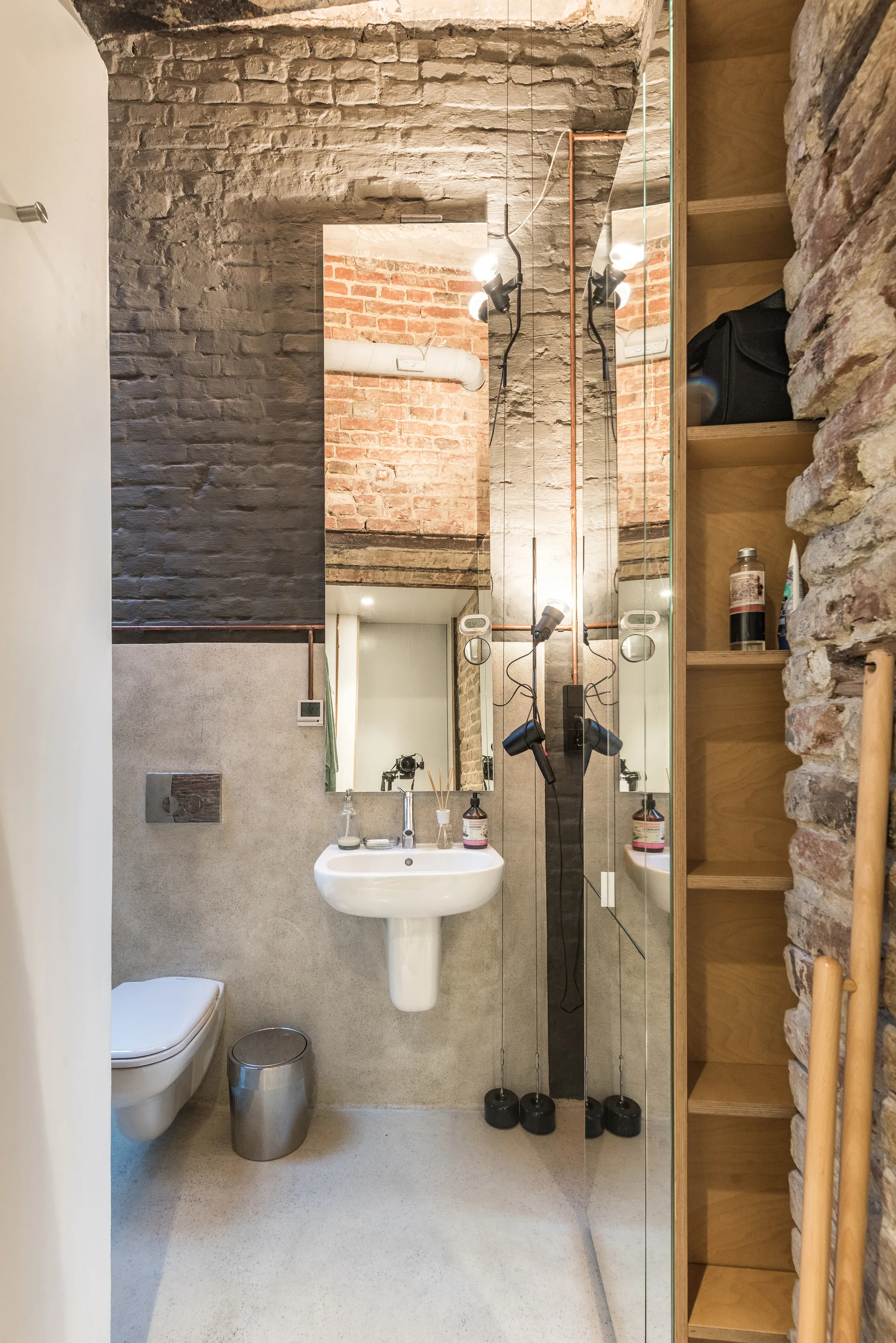 An industrial bathroom features a white pedestal sink, a wall-mounted toilet, exposed brick walls, and a tall wooden shelving unit.