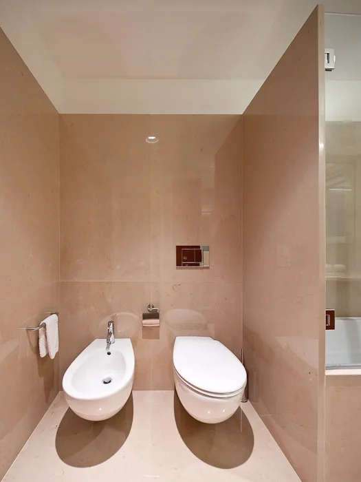 A minimalist bathroom featuring a wall-mounted white bidet and toilet against polished beige stone walls and matching tiled floors.
