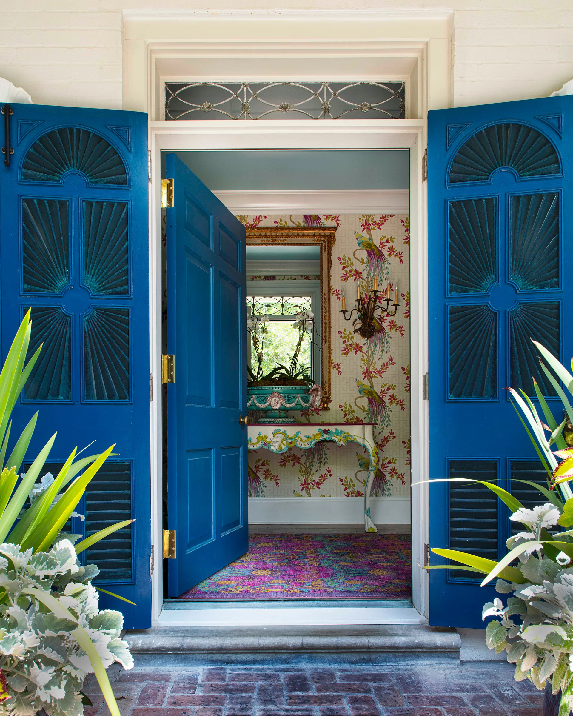 An open bright blue front door and matching shutters reveal an entryway decorated with floral wallpaper, an ornate console table, and a colorful rug.