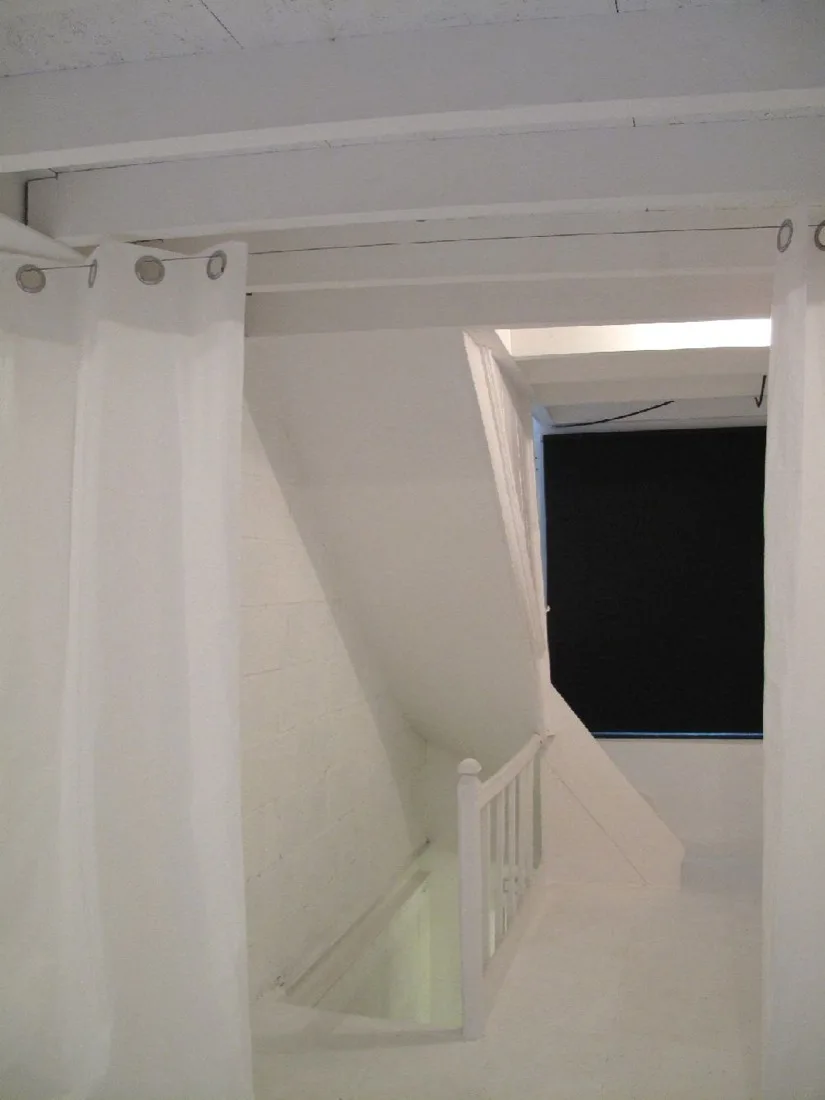 A minimalist white staircase features a wooden railing and exposed ceiling beams, framed by hanging white grommet curtains.