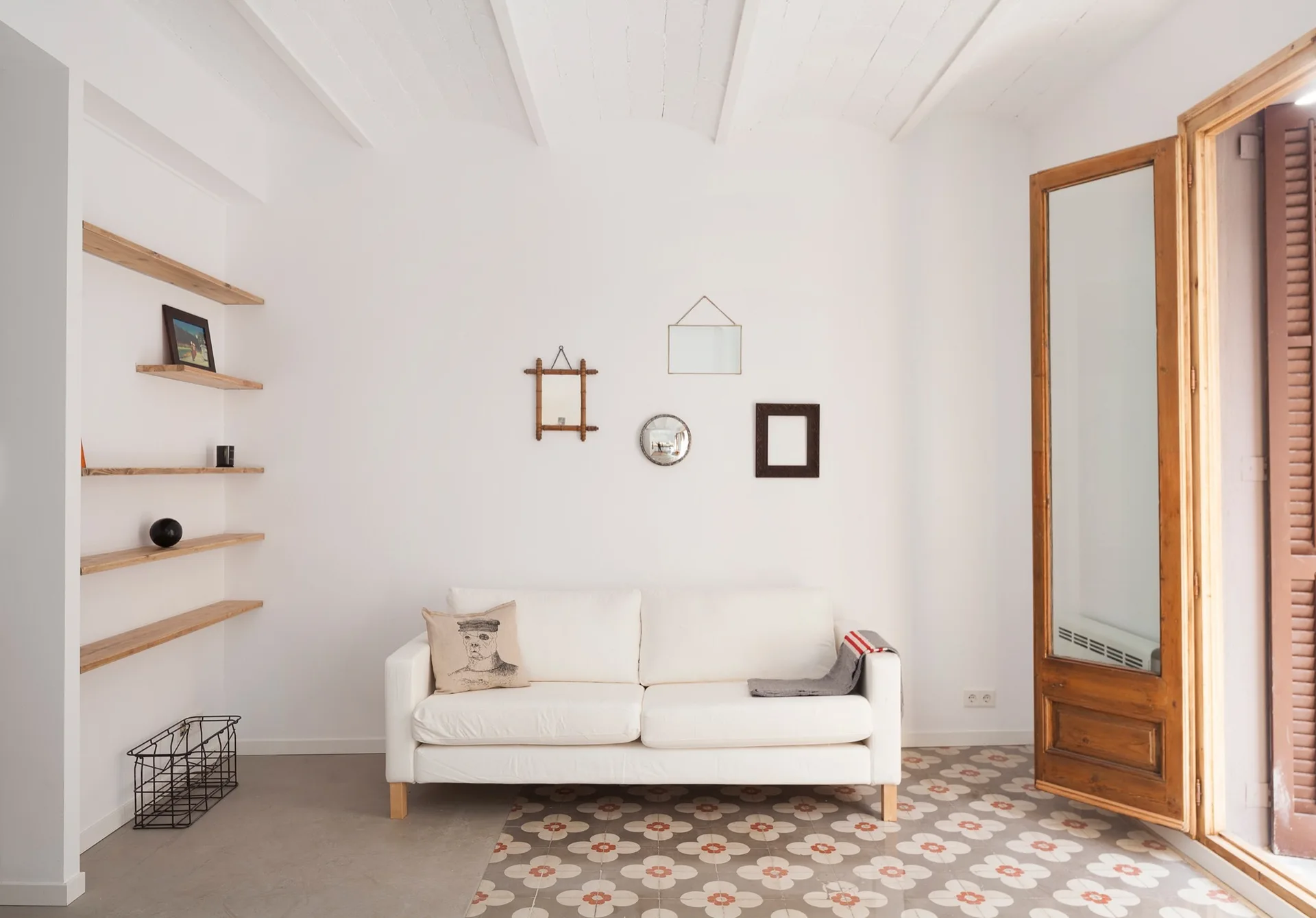 A minimalist white living room features a white sofa on patterned ceramic floor tiles beside floating wood shelves and a tall glass door.