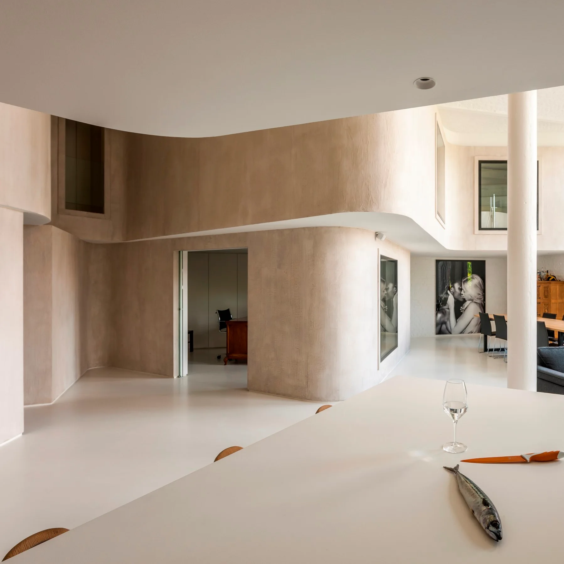A minimalist open-concept interior features smooth, curved beige plaster walls, a white floor, and a large white counter in the foreground.